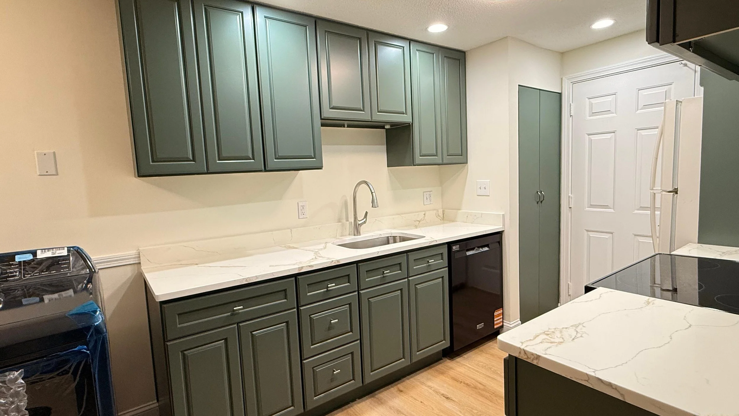A modern kitchen with green cabinetry, a white marble countertop, and a stainless steel sink. There are recessed ceiling lights and a white door in the background.