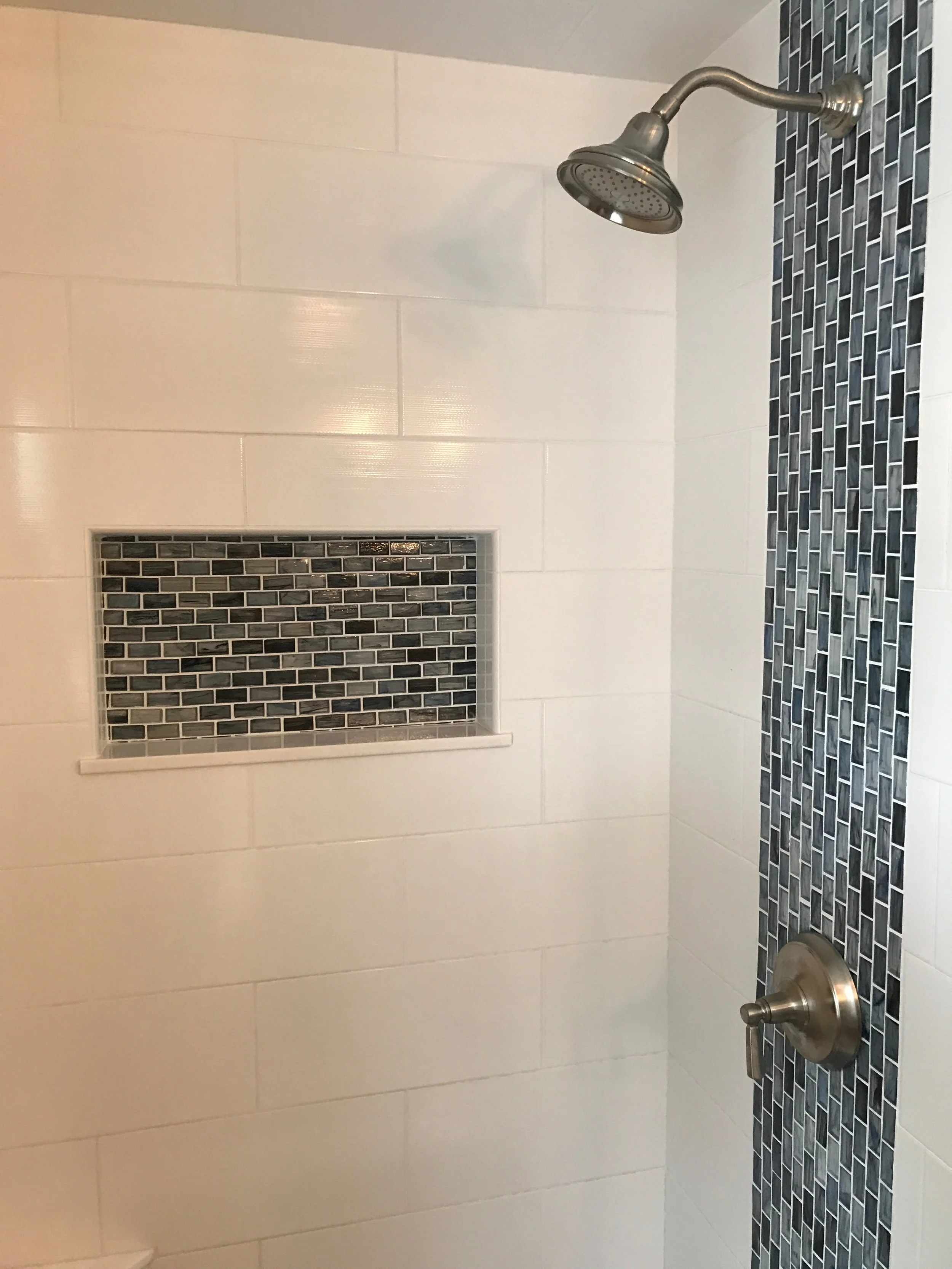 Bathroom shower with large white tiles on the wall, a small horizontal window with dark gray mosaic tiles, a chrome showerhead, and a matching chrome water control knob.