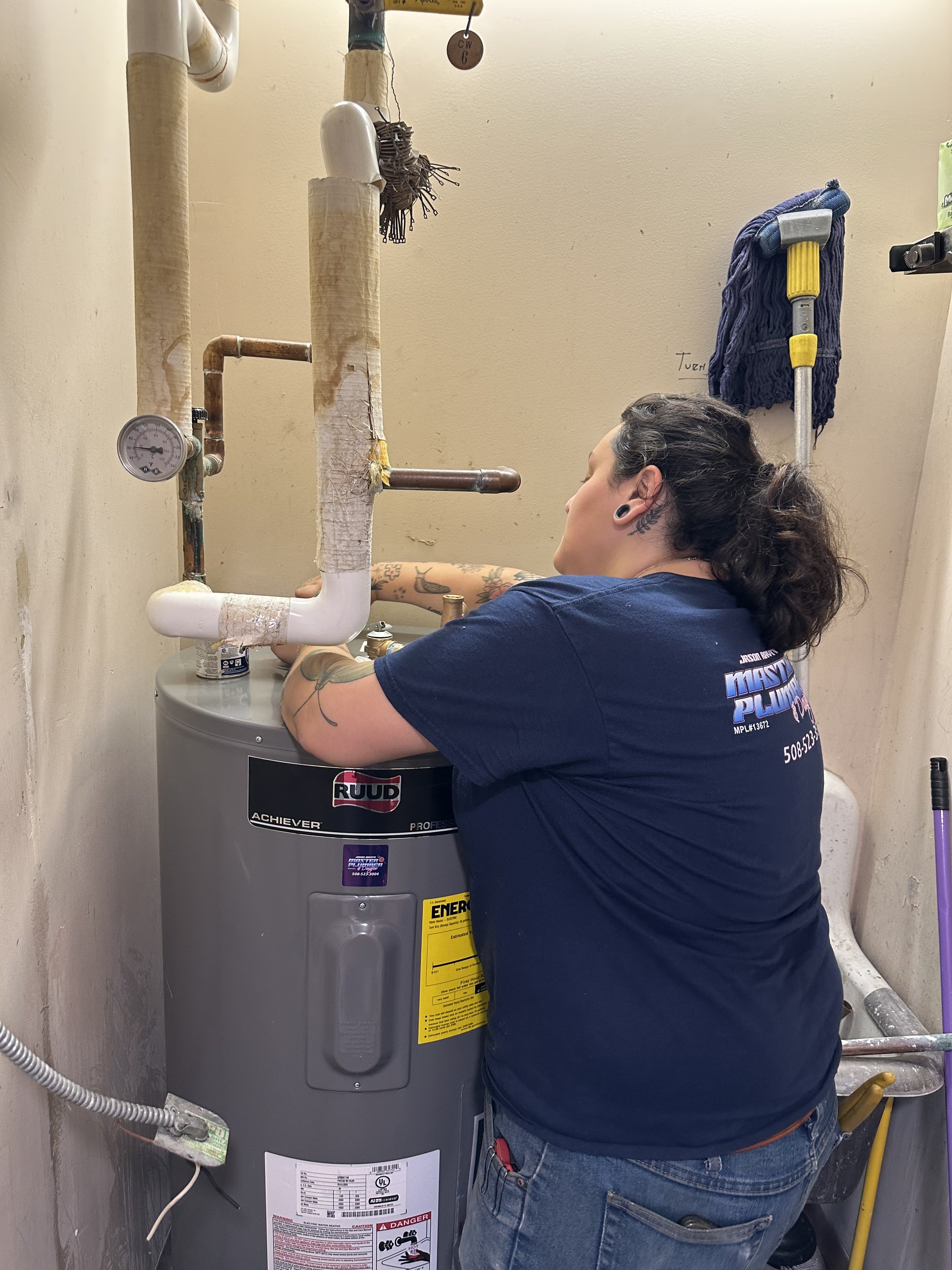 A person with tattoos on their arm working on a water heater in a small utility room.