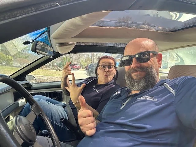 A man and woman taking a selfie inside a car. The man has a beard, is wearing sunglasses and a blue shirt, and is giving a thumbs-up. The woman is making a surprised or playful face and wearing glasses.