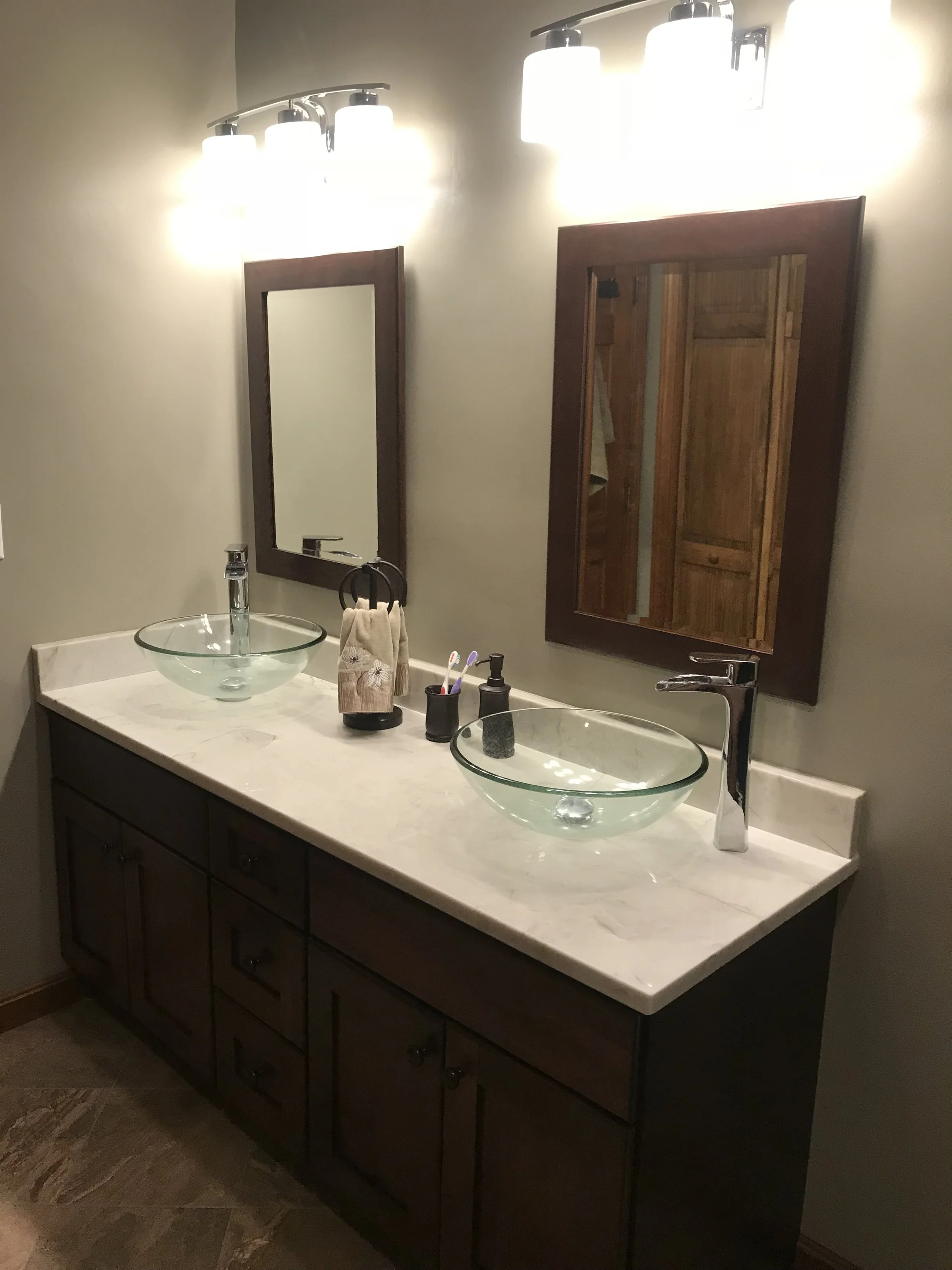 Bathroom double vanity with dark wooden cabinets, two round glass vessel sinks, modern chrome faucets, two rectangular mirrors with dark wooden frames, overhead light fixtures, and bathroom essentials like toothpaste and soap.