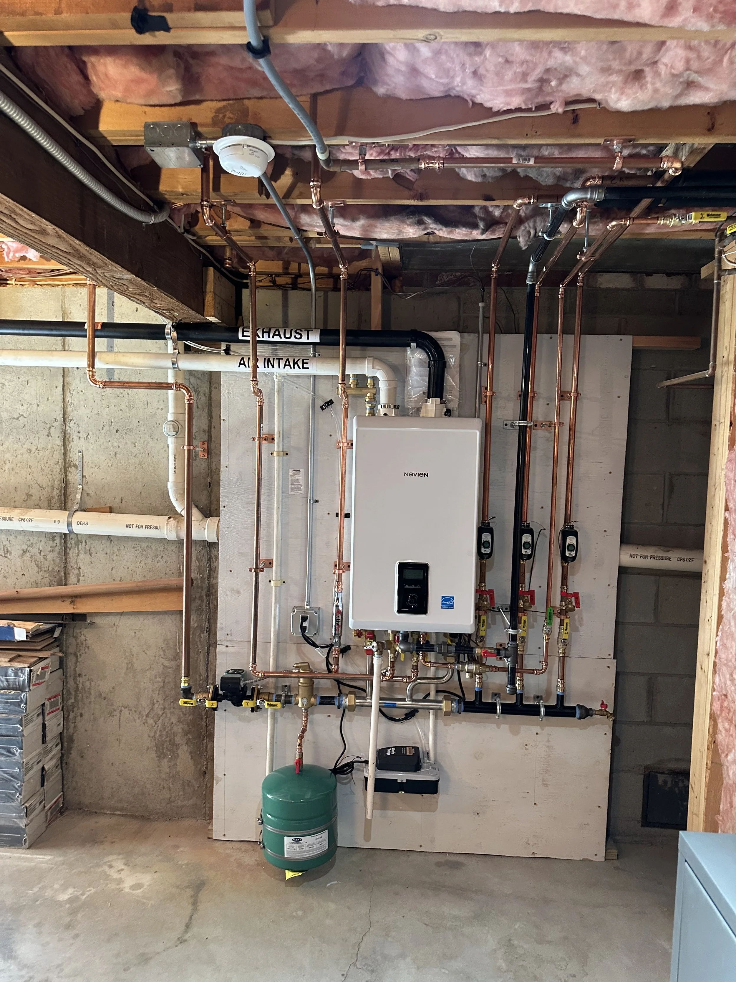 A basement with plumbing and HVAC system including copper pipes, valves, a green expansion tank, and a Navien tankless water heater mounted on a plywood wall, with insulation on the ceiling and concrete floor.
