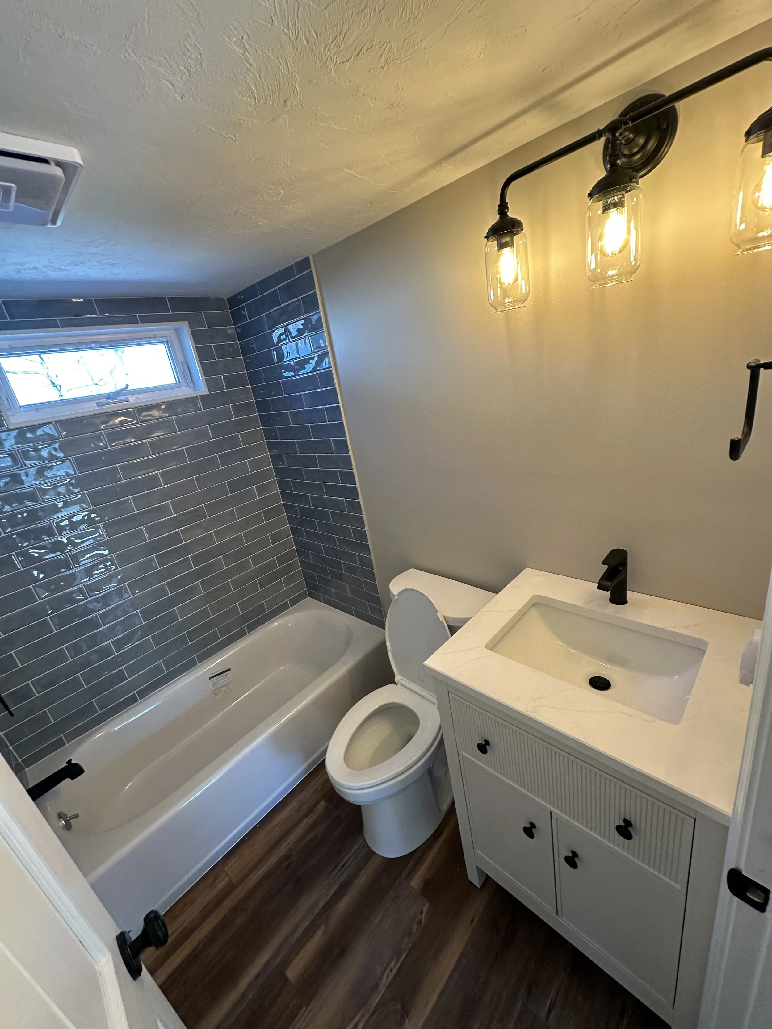 A small bathroom with a bathtub, a toilet, a white vanity with a sink, black faucet, and a mirror. The wall behind the sink is painted light, and the wall behind the bathtub has dark gray subway tiles. There is a small window in the corner and a mode