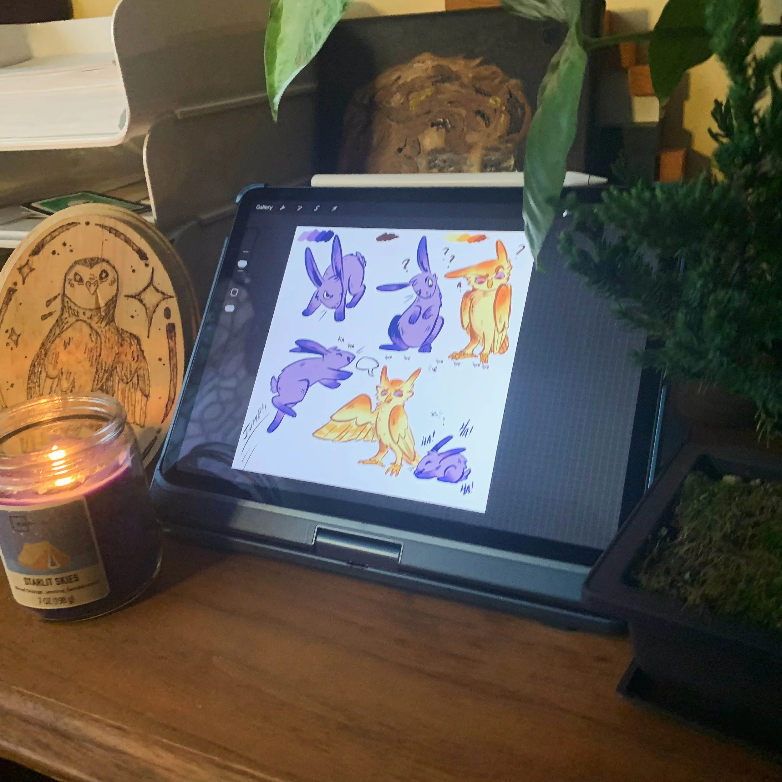 Tablet on a desk displaying character sketches of a bunny and an owl in different poses and expressions, with plants, books, a lit candle, and decorative wooden art pieces surrounding it.