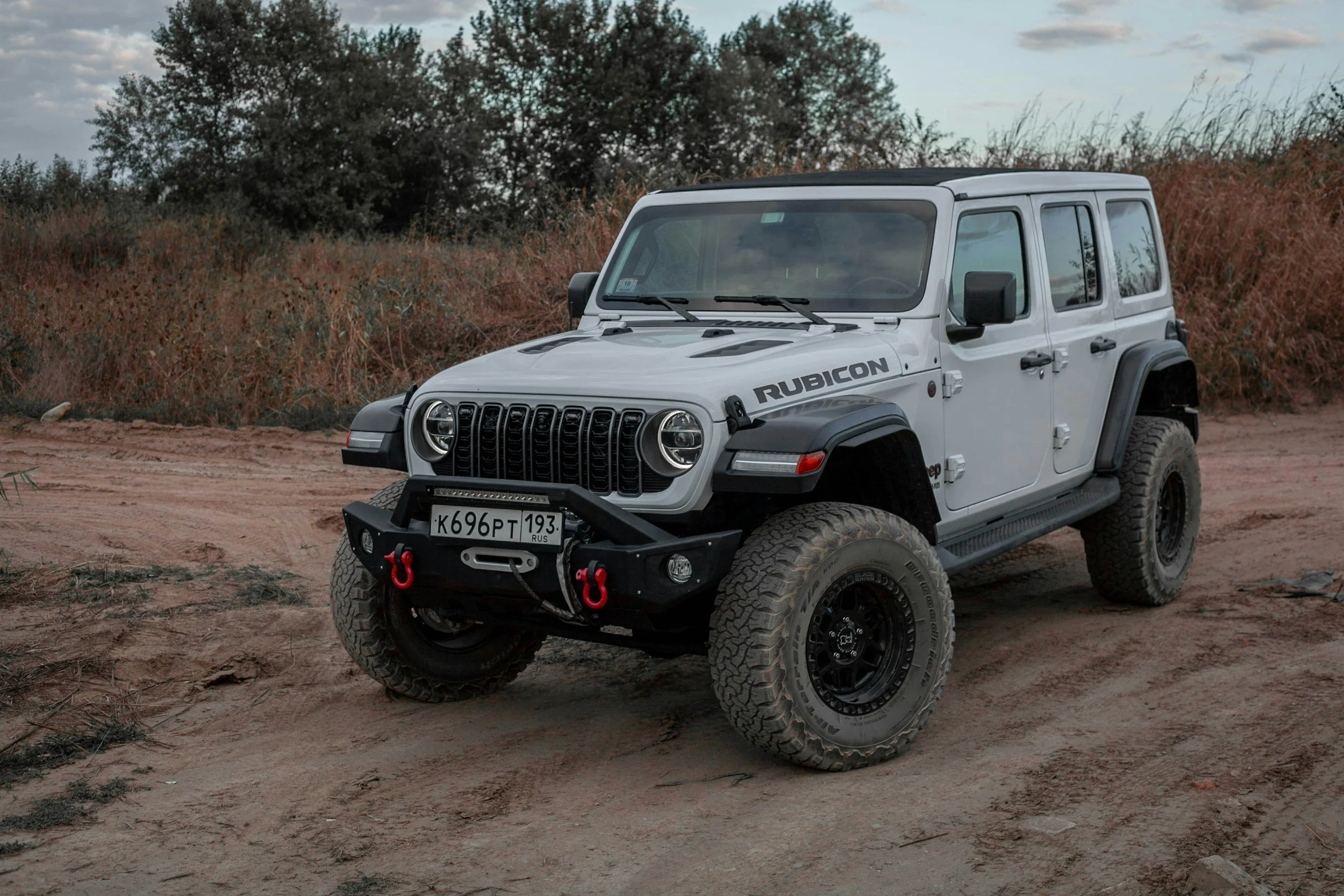 A white Jeep Rubicon off-road vehicle with black wheels, a front bumper with red hooks, and a Russian license plate, parked on dirt with bushes and trees in the background.