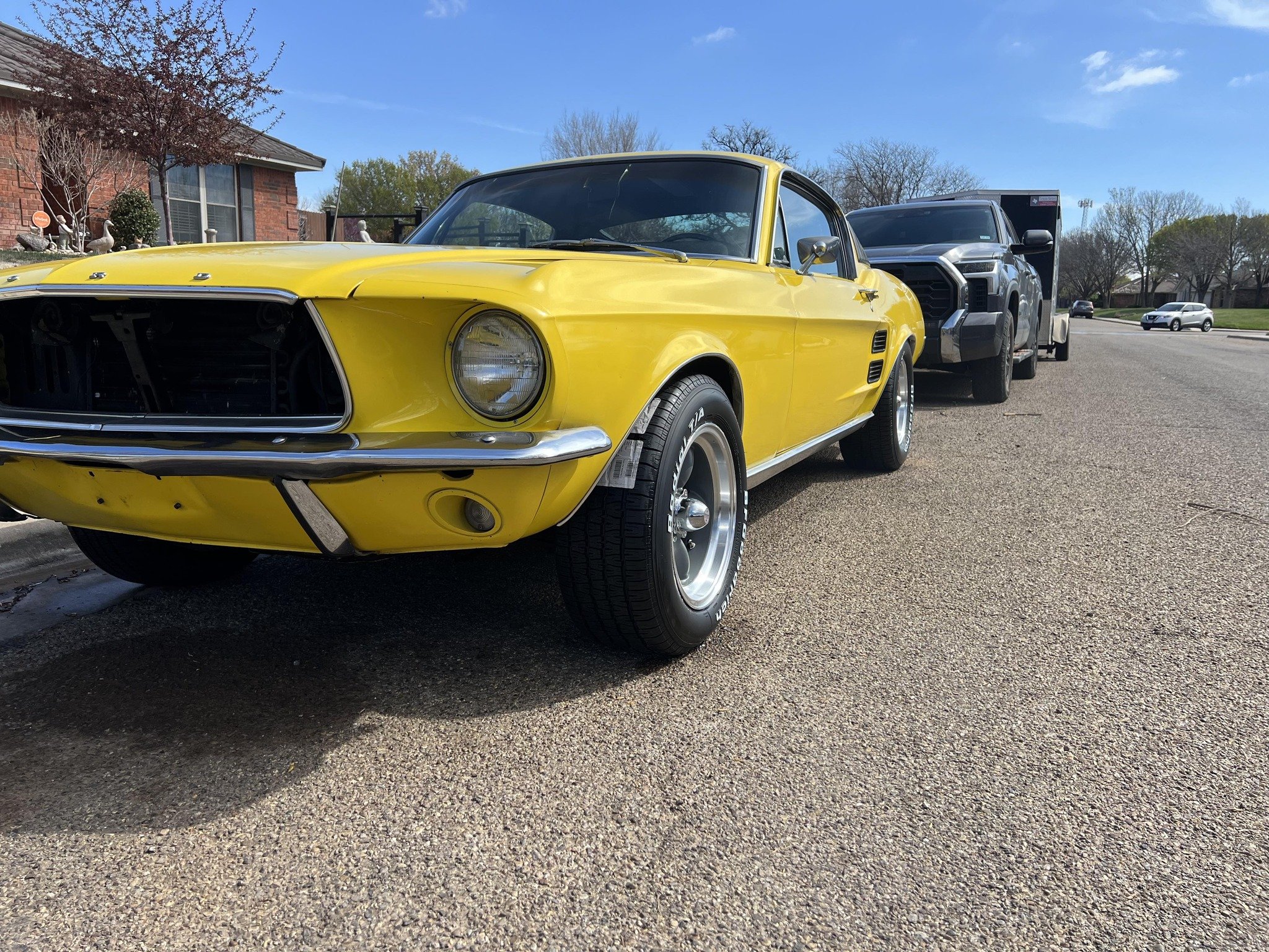 Yellow classic Ford Mustang car parked on the side of the road with a modern black pickup truck behind it, under a blue sky with some clouds.