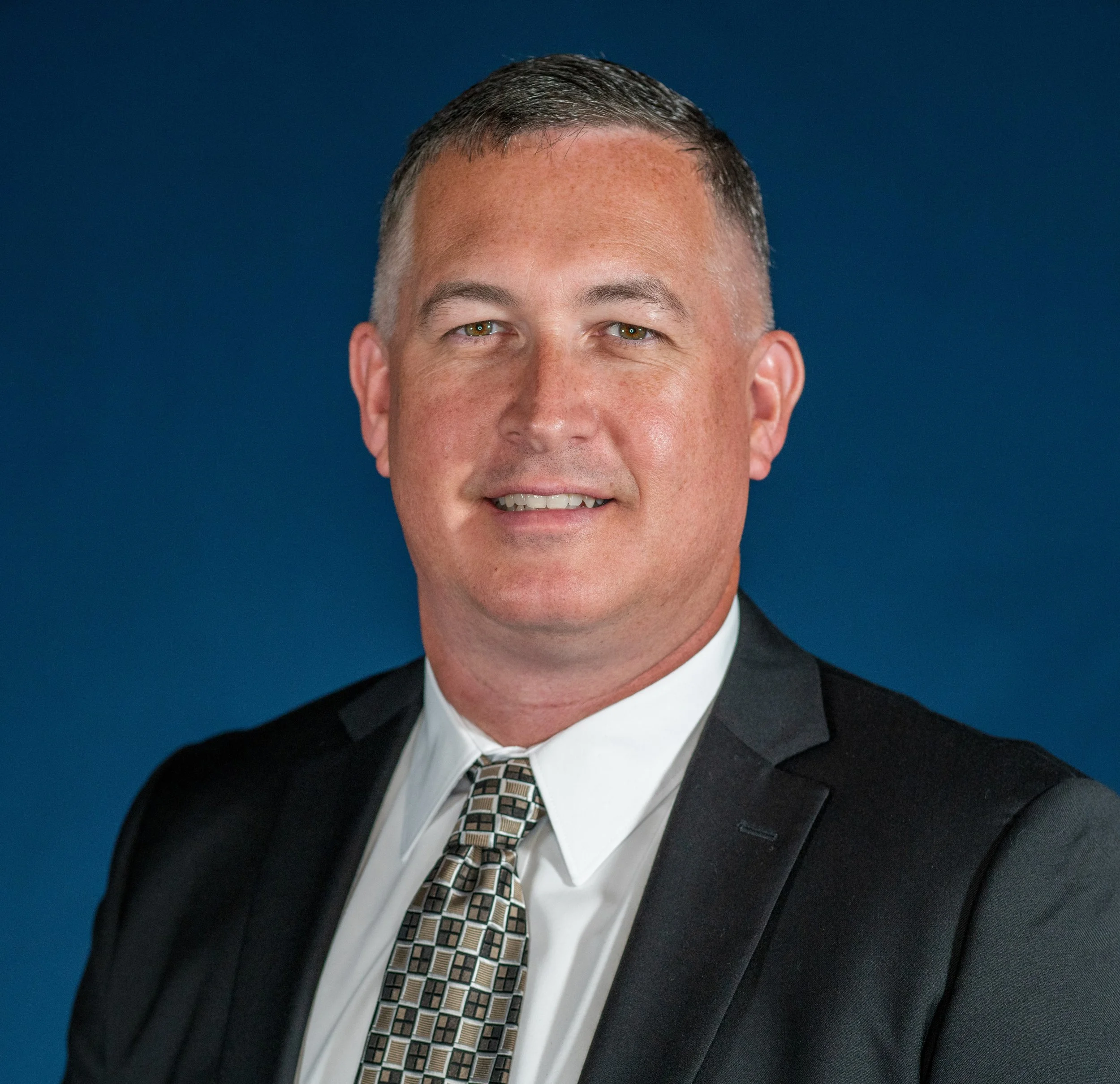 Portrait of a middle-aged man in a black suit, white shirt, and patterned tie, smiling against a dark blue background.