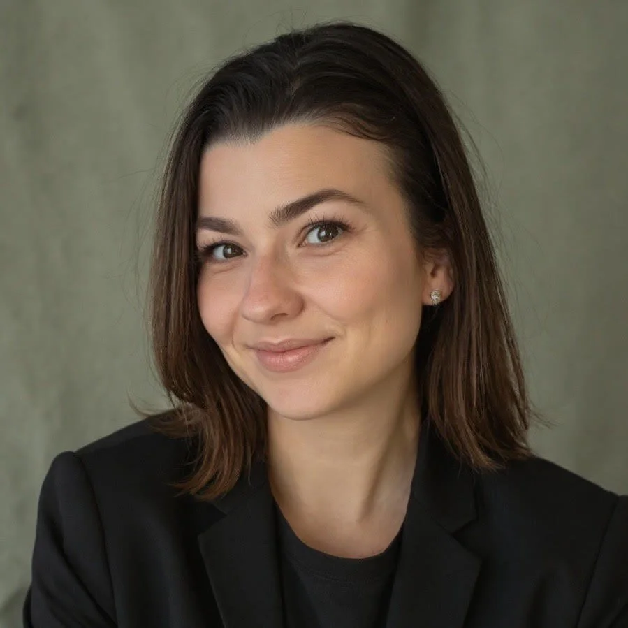 A woman with shoulder-length brown hair, wearing a black blazer and earrings, smiling softly against a neutral background.