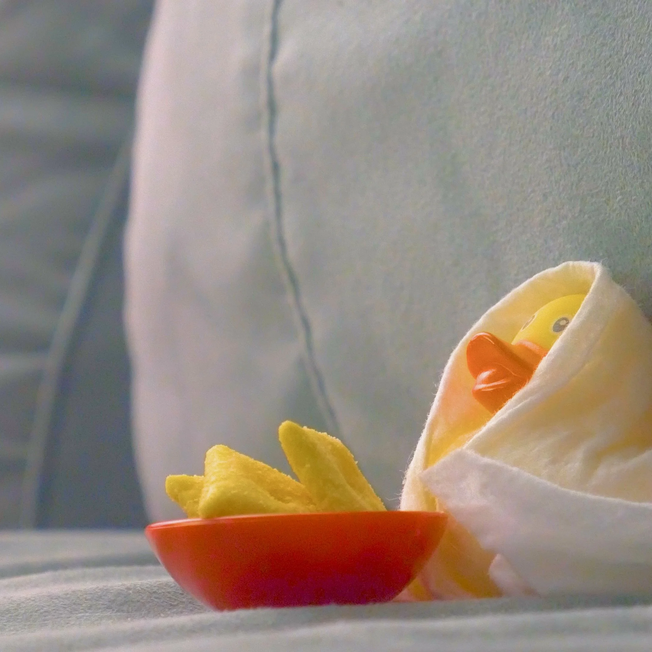 A yellow rubber duck with an orange beak inside a white cloth napkin, next to a small red bowl of potato chips, on a cotton surface.