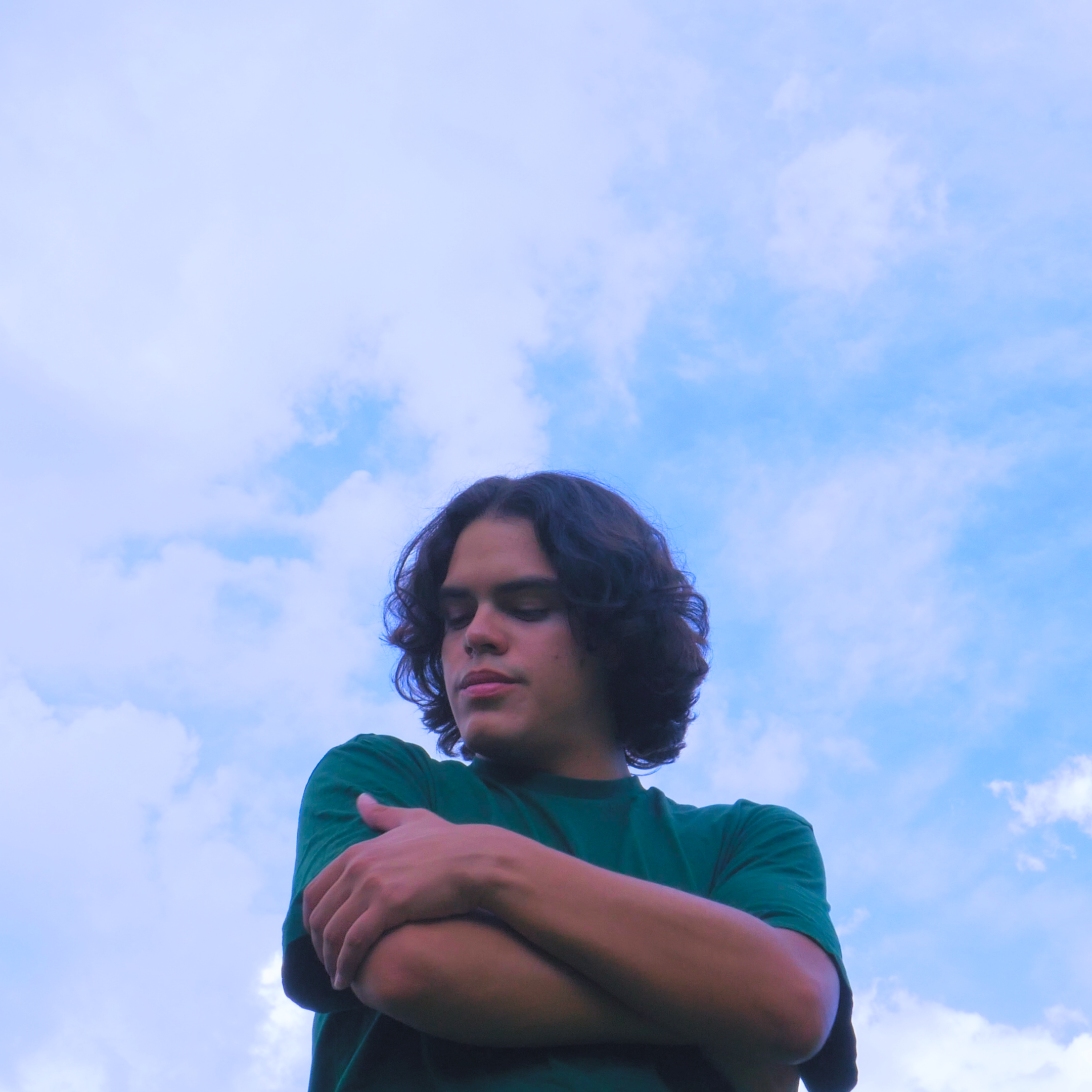 A young man with long curly hair standing outdoors against a background of blue sky and clouds, with his arms crossed over his chest and eyes closed.
