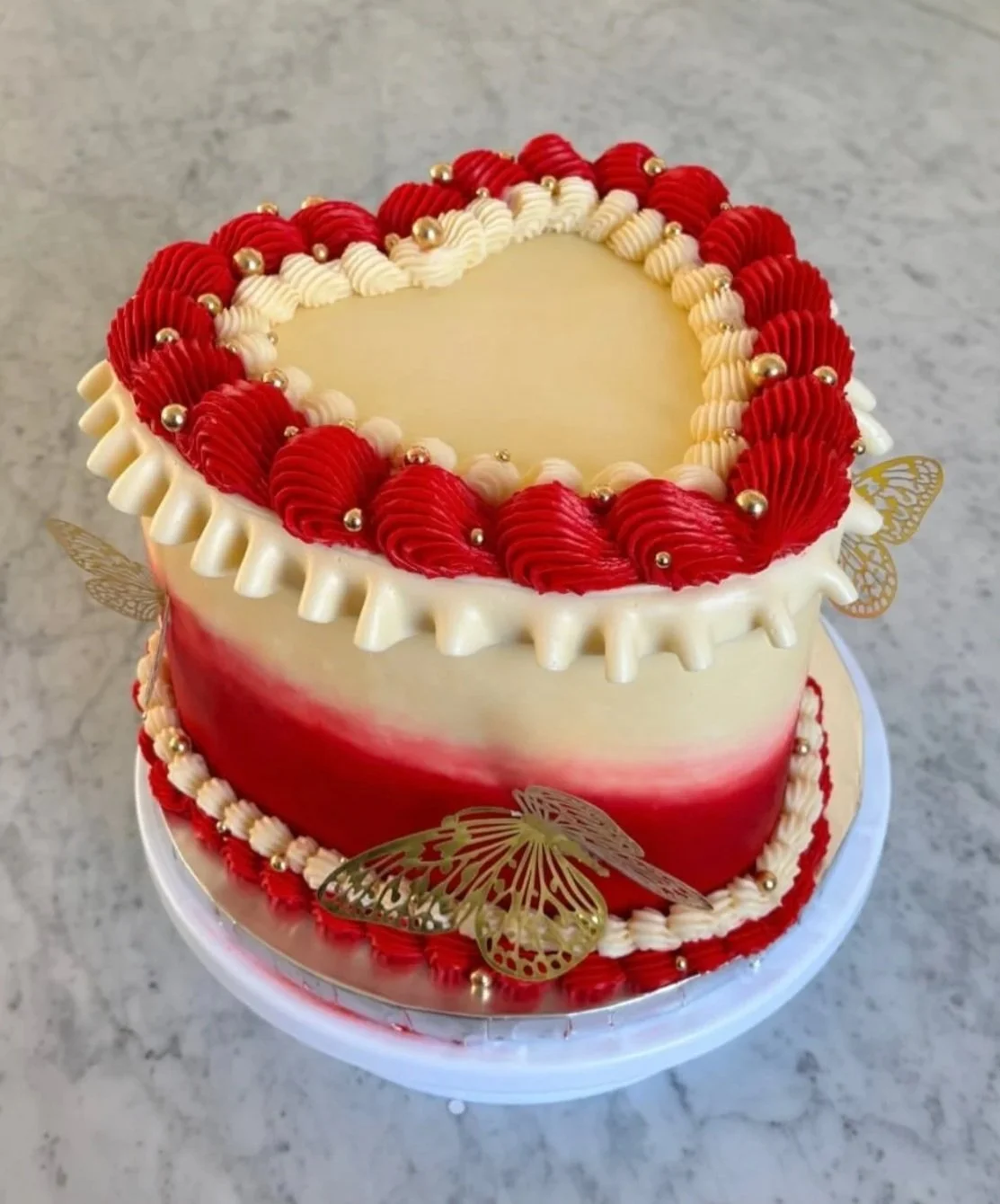  Scarlet Romance    
A charming red and white heart-shaped cake layered with chocolate sponge and vanilla buttercream, topped with hand-piped rosettes and gold butterflies — a timeless symbol of love and celebration. 