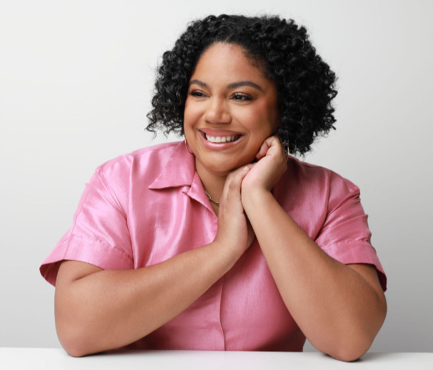 Image of Lauren Carson,a woman with curly hair smiling and resting her face on her hands, wearing a pink satin shirt against a neutral background.