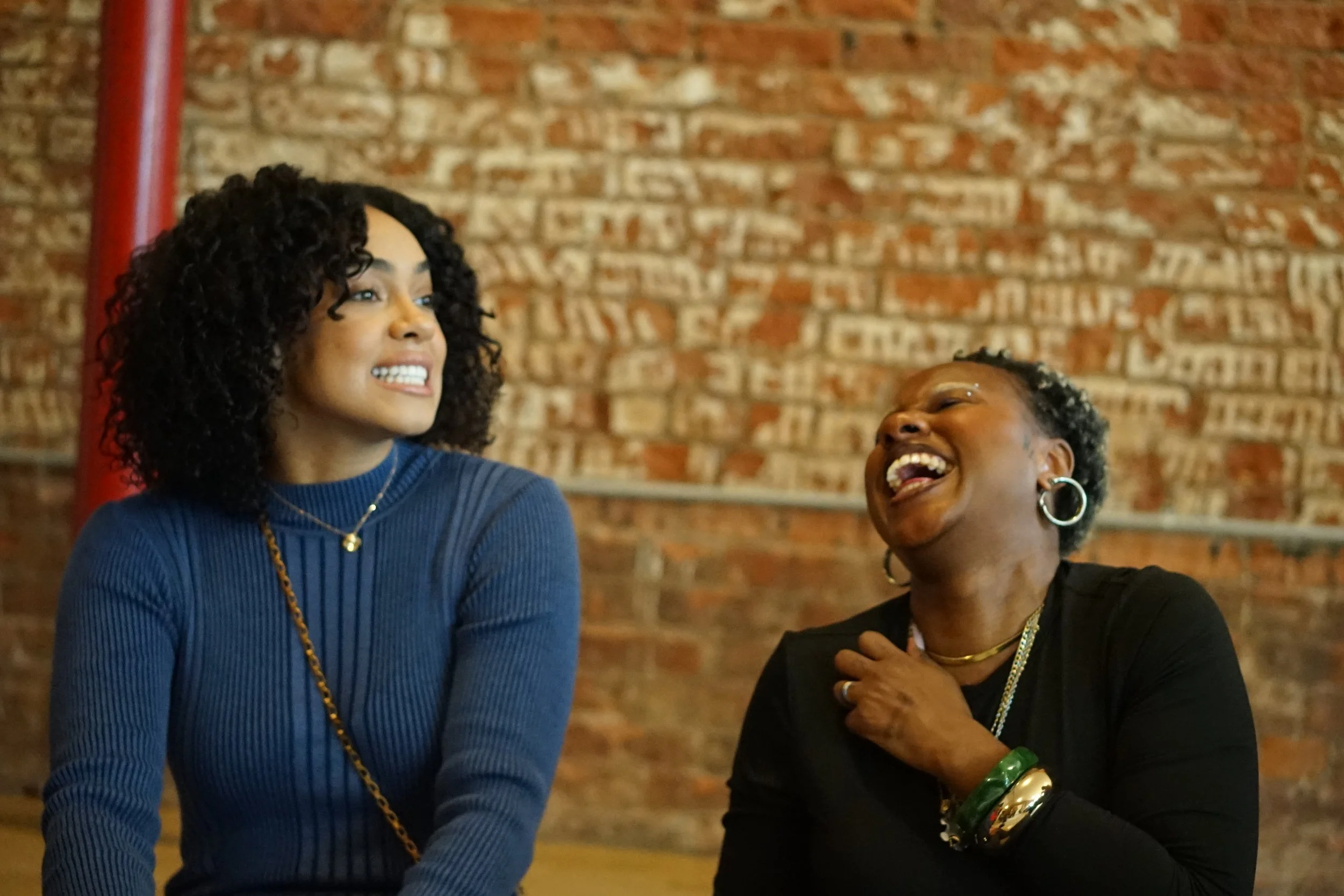 Two women are laughing and smiling at each other in front of a brick wall at a Black Girls Smile event. One has curly black hair and is wearing a blue top, the other has short curly hair, wearing large earrings, and a black top with jewelry.