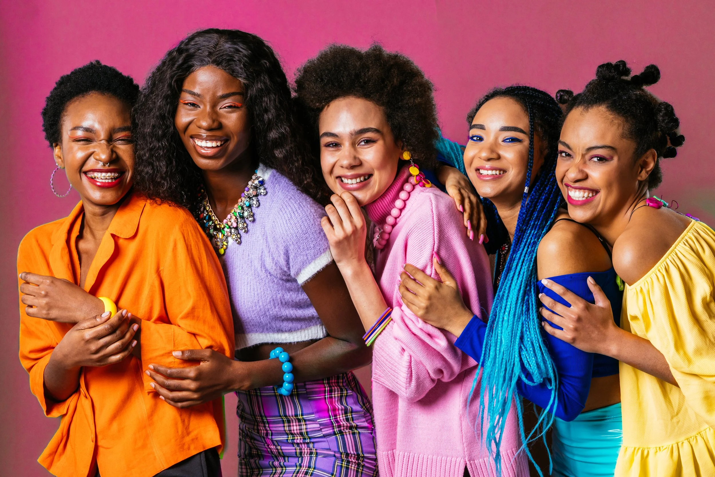 Six women of diverse styles smiling and hugging against a pink background, dressed in bright colorful clothes and accessories.