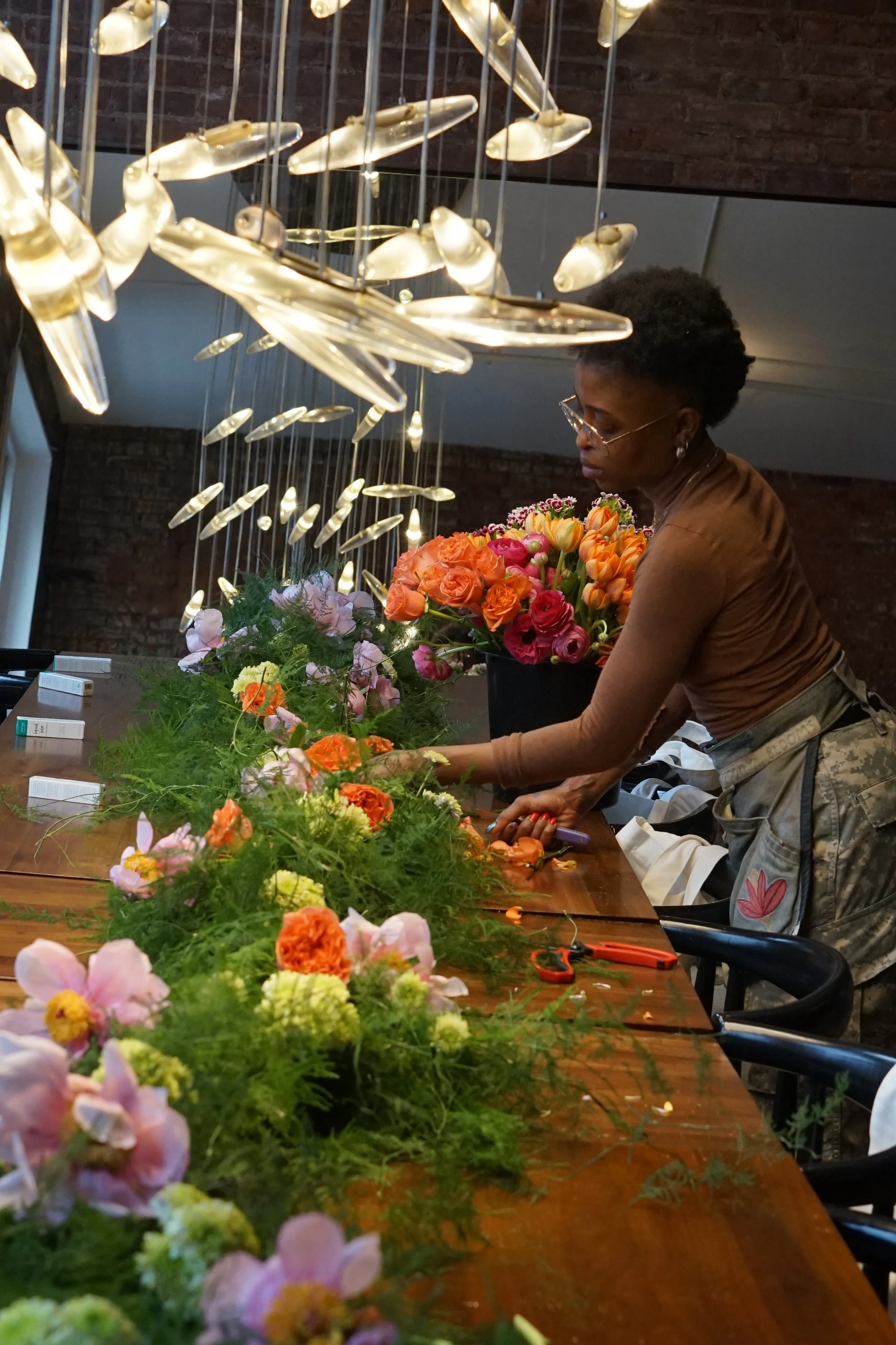 A woman arranging pink, orange, and yellow flowers on a long wooden table, with a lush green floral arrangement in front of her. She is wearing glasses, a brown top, and camouflage pants, inside a room with an artistic light fixture hanging from the ceiling.
