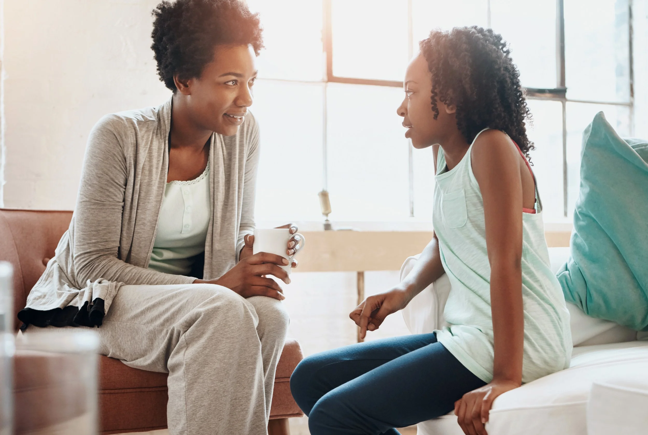 A woman and a girl sitting and talking in a bright, sunlit room, with the woman holding a white mug.
