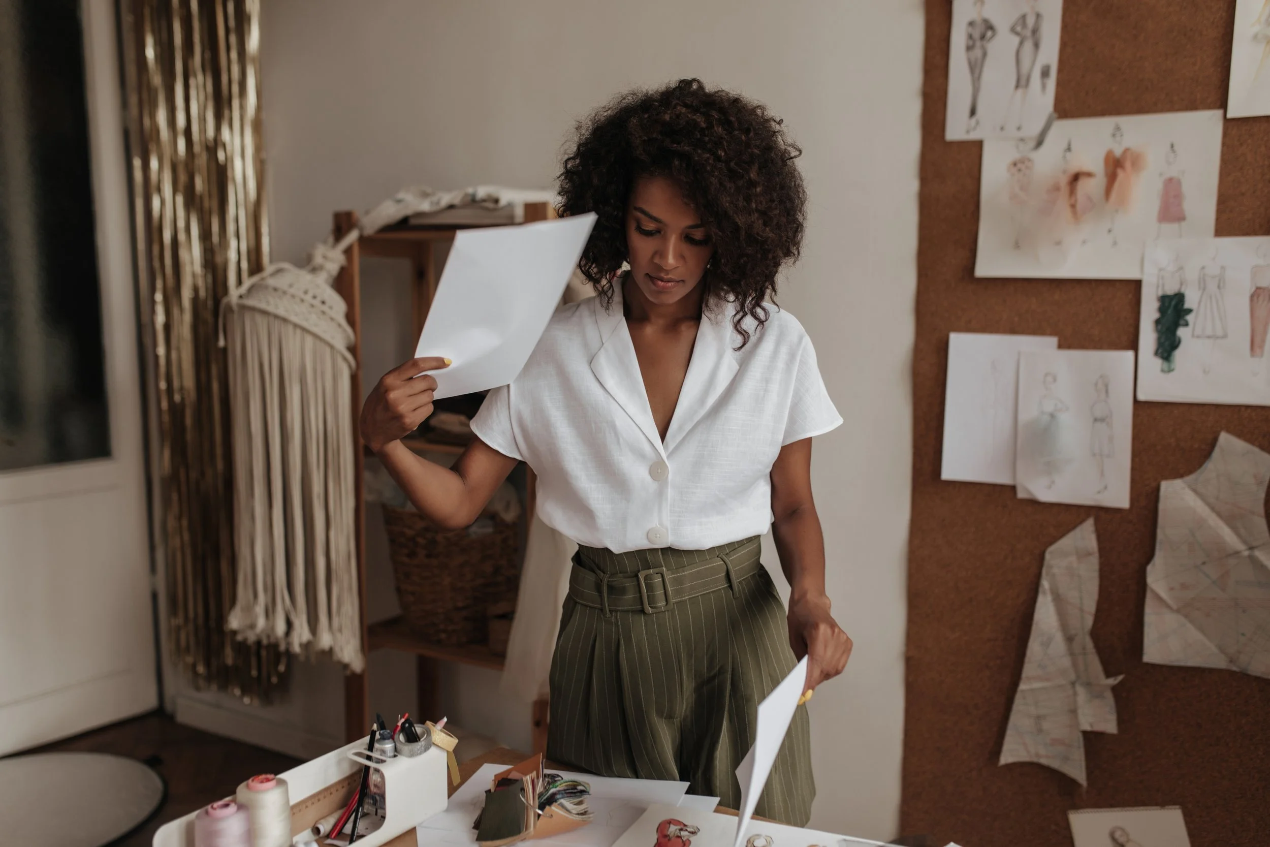 A woman with curly hair wearing a white short-sleeve blouse and high-waisted green pinstripe pants in a workspace with sketches on a corkboard and drawing supplies on the table.