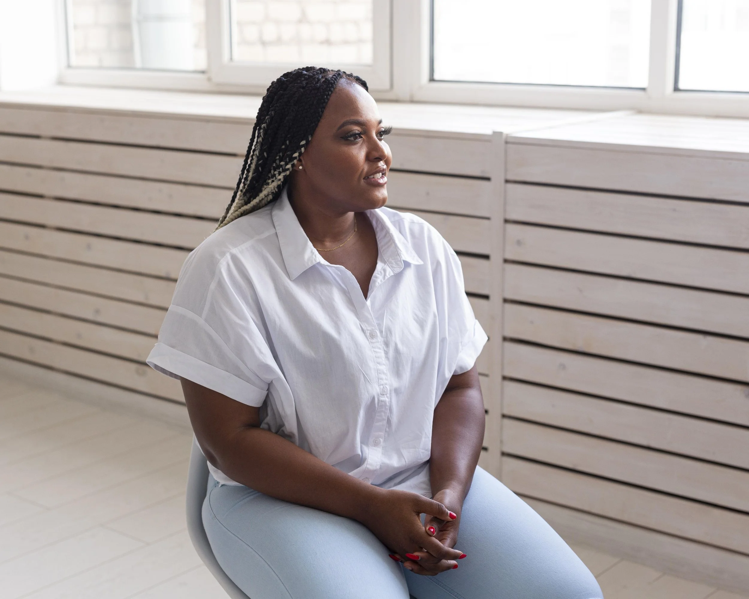 A woman with braided hair sitting and looking thoughtfully in a room with large windows and wooden paneling