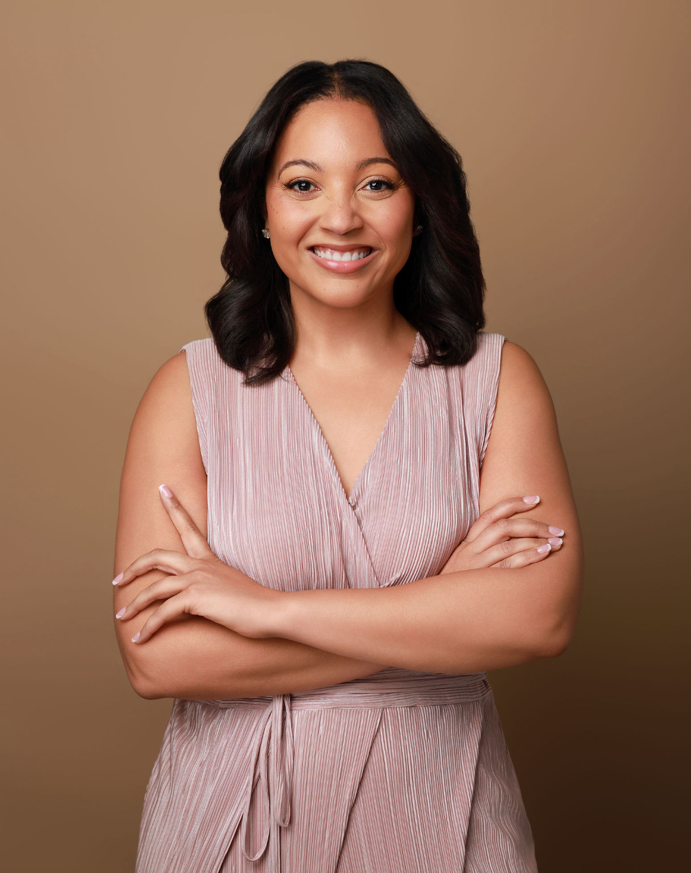 A smiling woman with medium-length black hair, wearing a sleeveless pink dress, crossed her arms, and standing against a beige background.