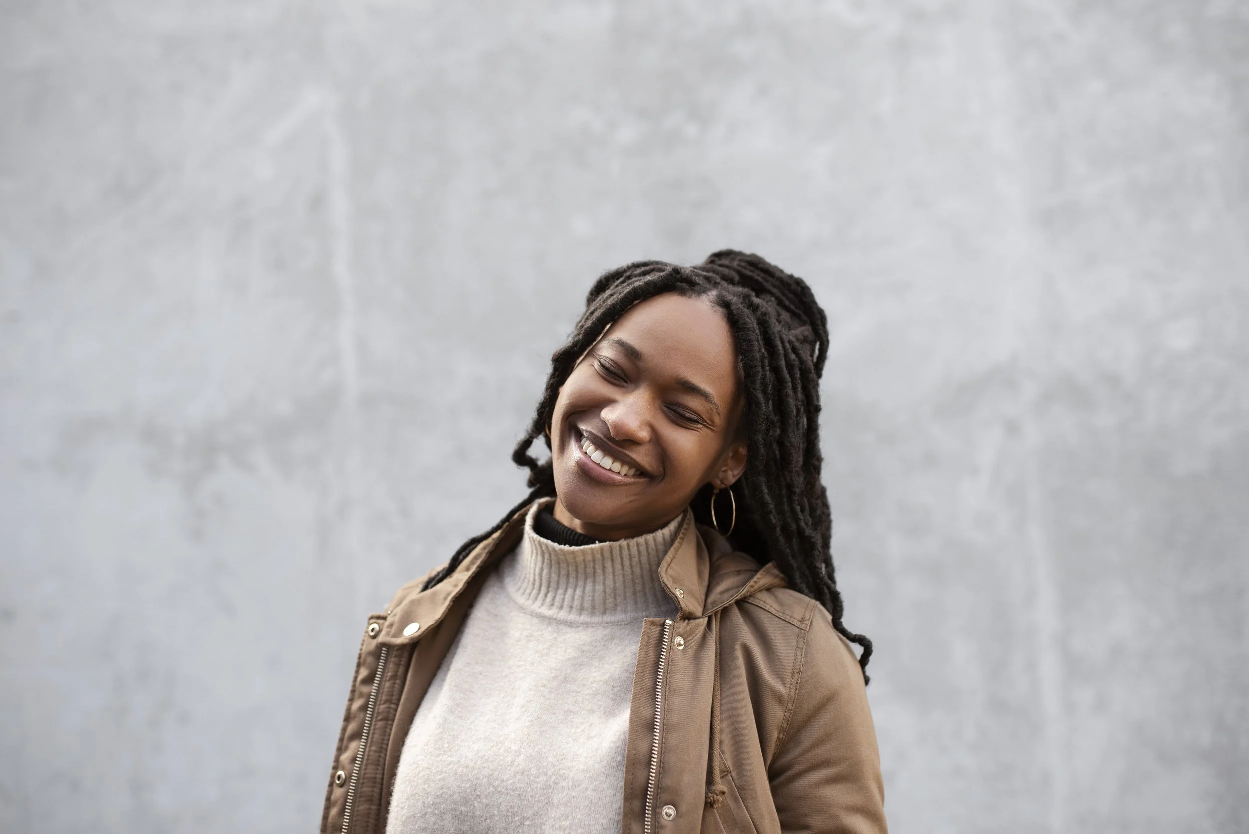 A young woman with long dreadlocks smiling with eyes closed, standing against a plain gray wall, wearing a beige sweater and a brown jacket.