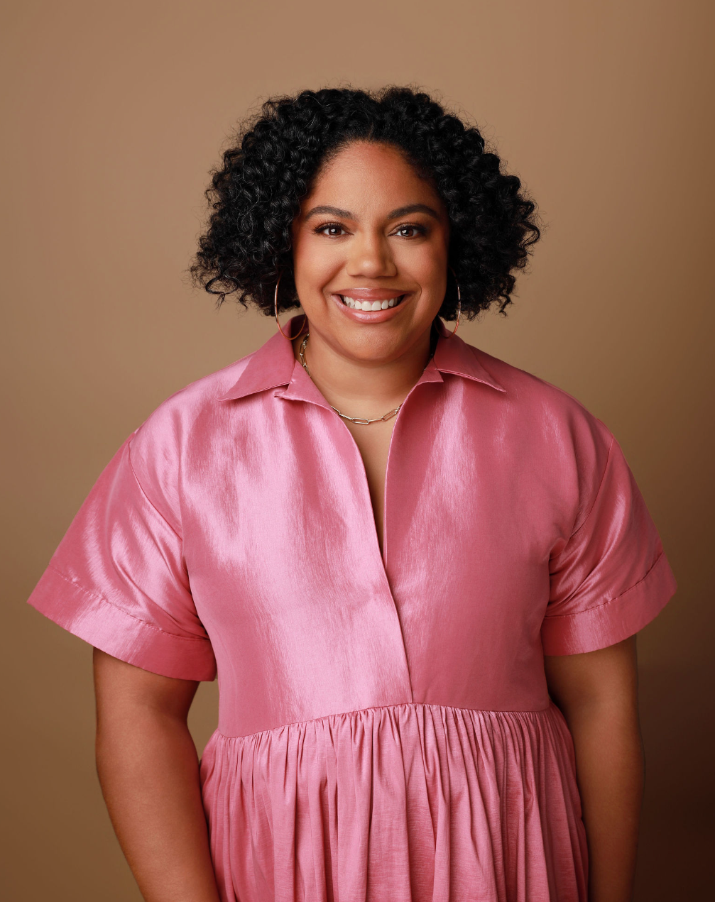 Headshot of a woman with dark curly hair, smiling, wearing a pink satin dress, hoop earrings, and a necklace, against a beige background.