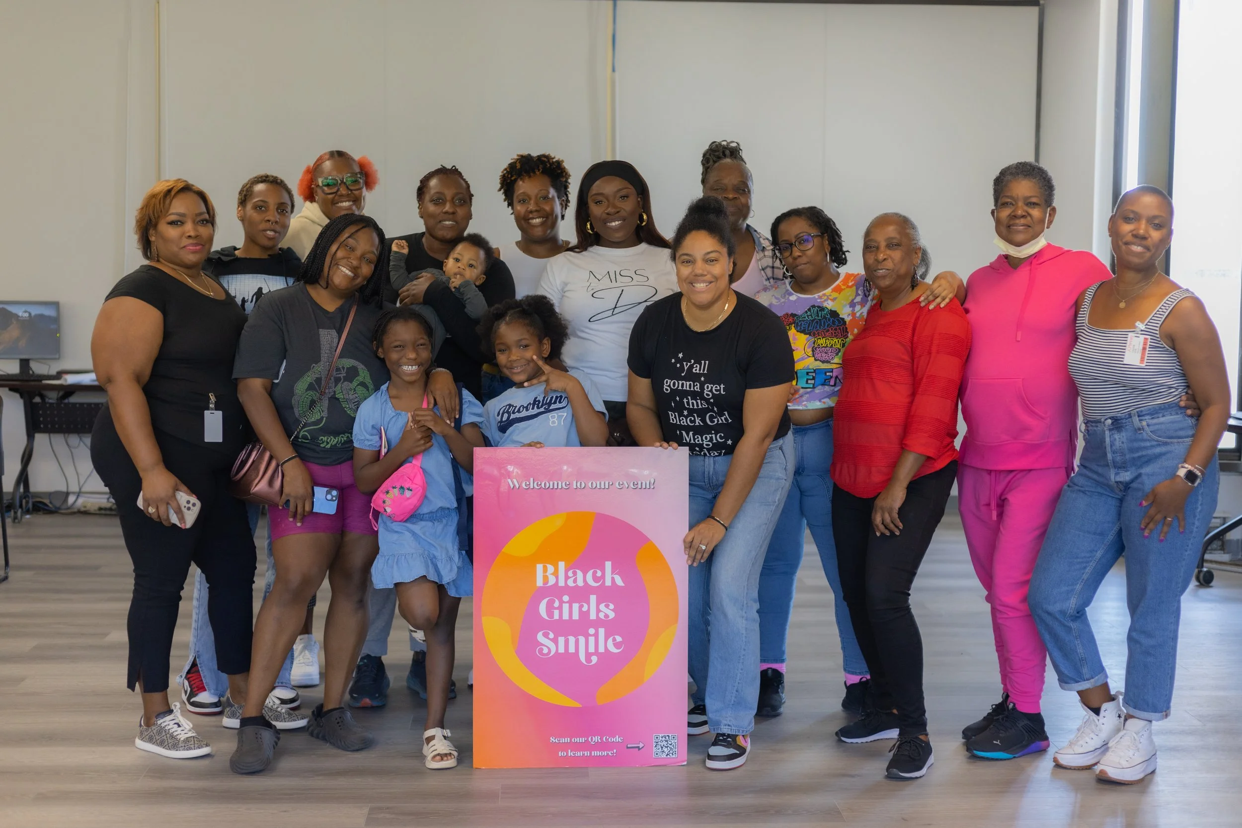 Group of women and children smiling for a photo at an indoor event with a pink sign that reads "Black Girls Smile".