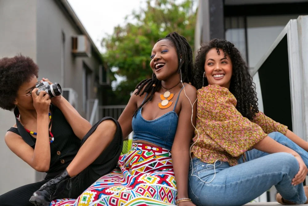 Three young women sitting outdoors, smiling and enjoying music, with one taking a photo.