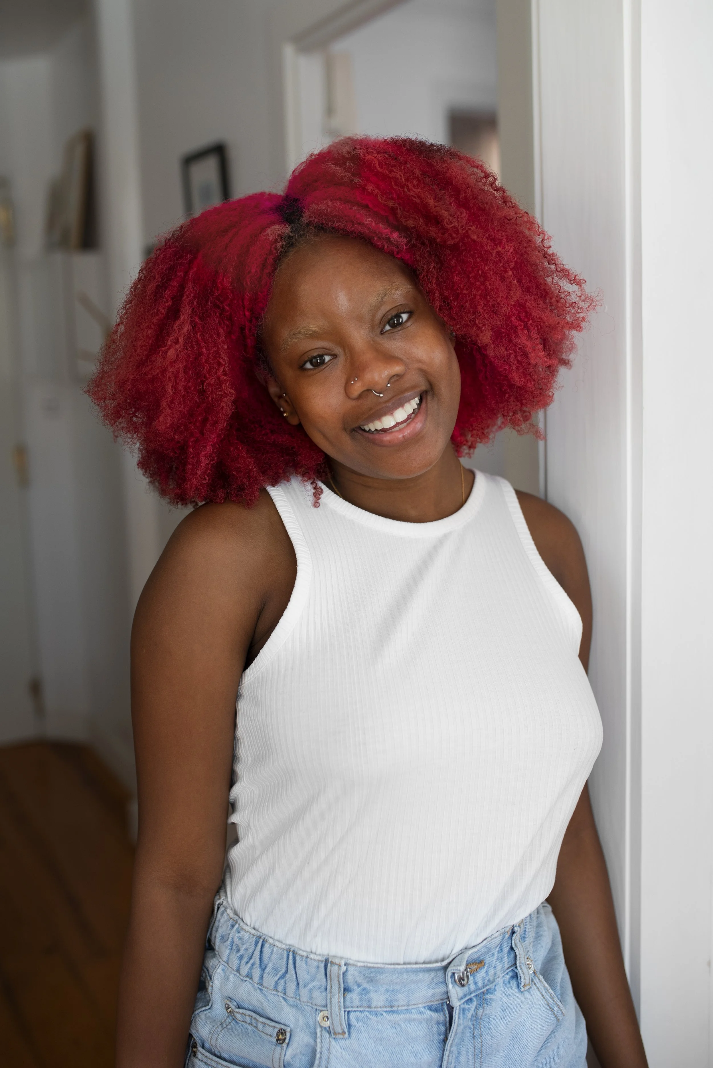 A woman with vibrant red curly hair smiling while leaning against a doorway in a home interior.