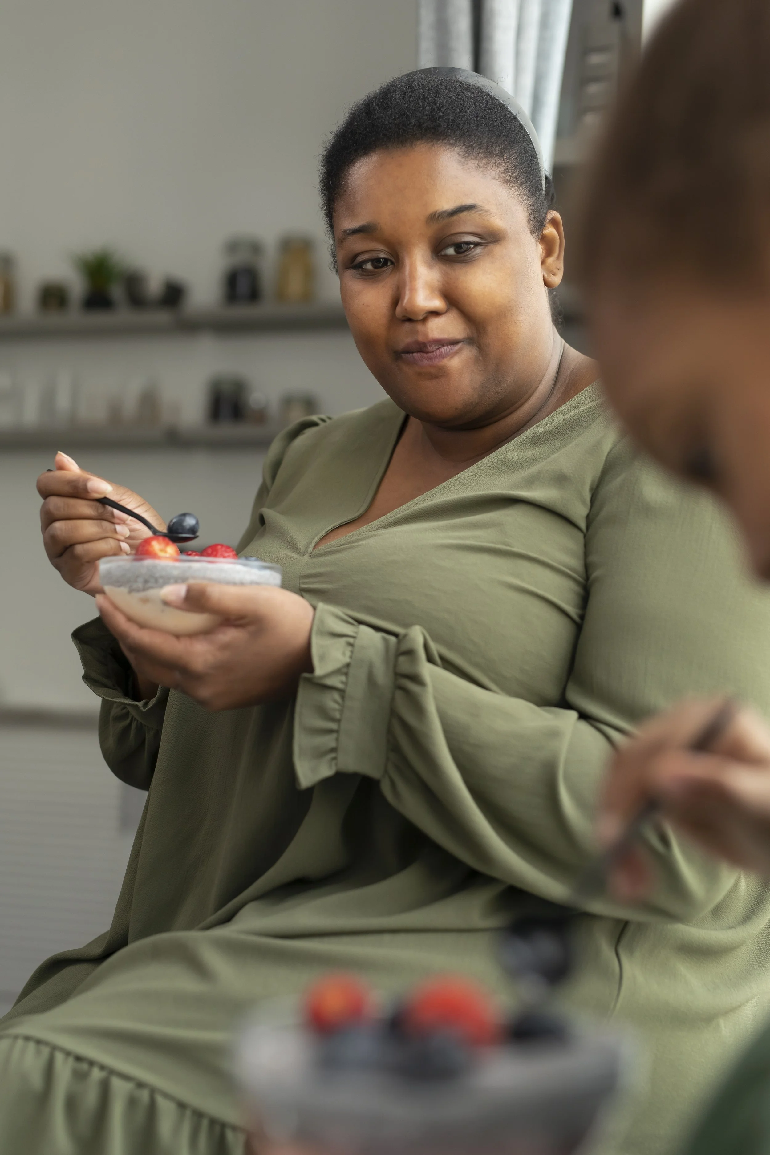 A woman in a green dress holding a bowl of pudding with berries, sitting indoors.
