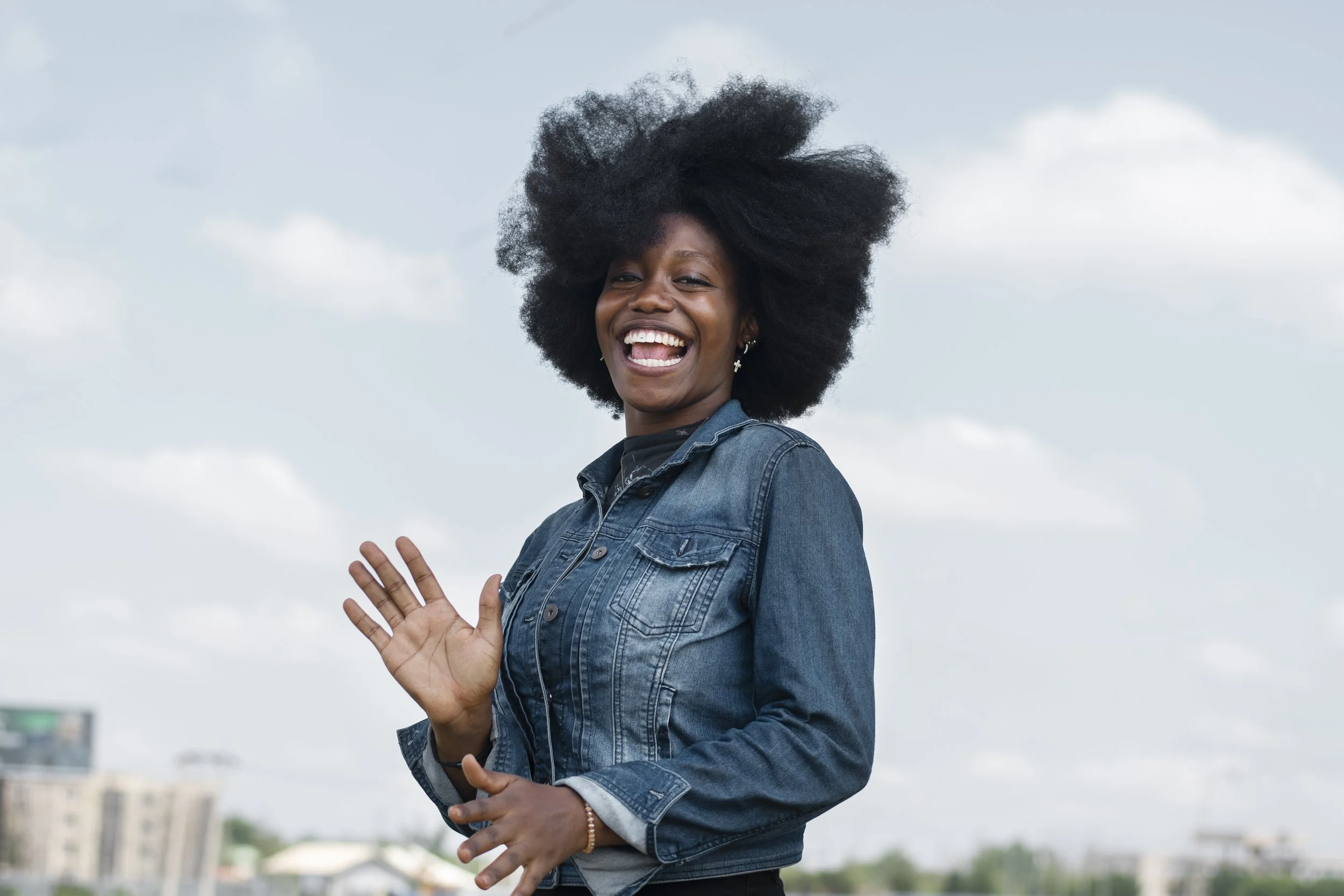 A woman with large curly black hair smiling and waving outdoors on a cloudy day