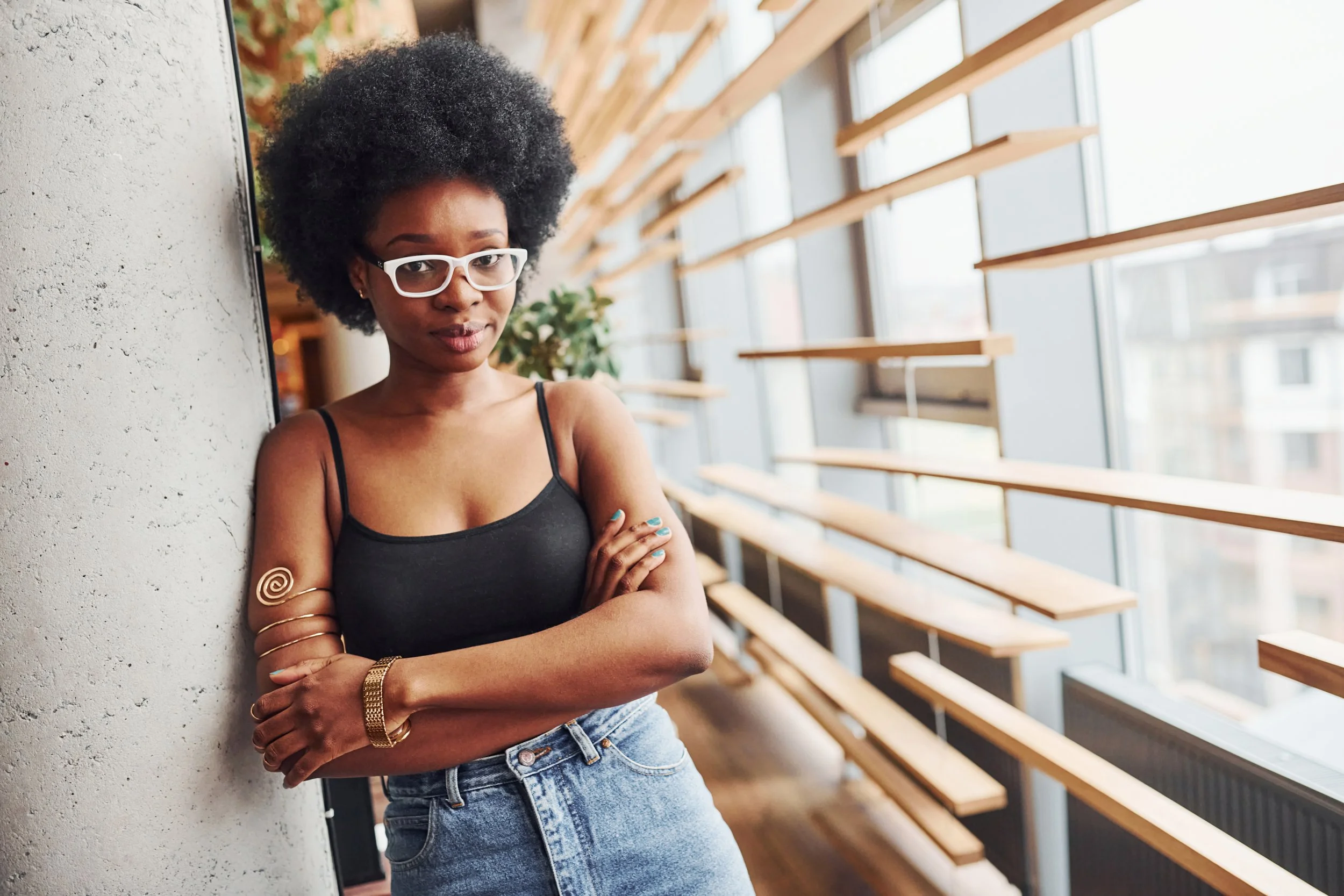 A young woman with curly black hair, wearing white glasses, a black tank top, light denim jeans, and gold accessories, standing indoors near a concrete wall, with large windows and wooden benches in the background.
