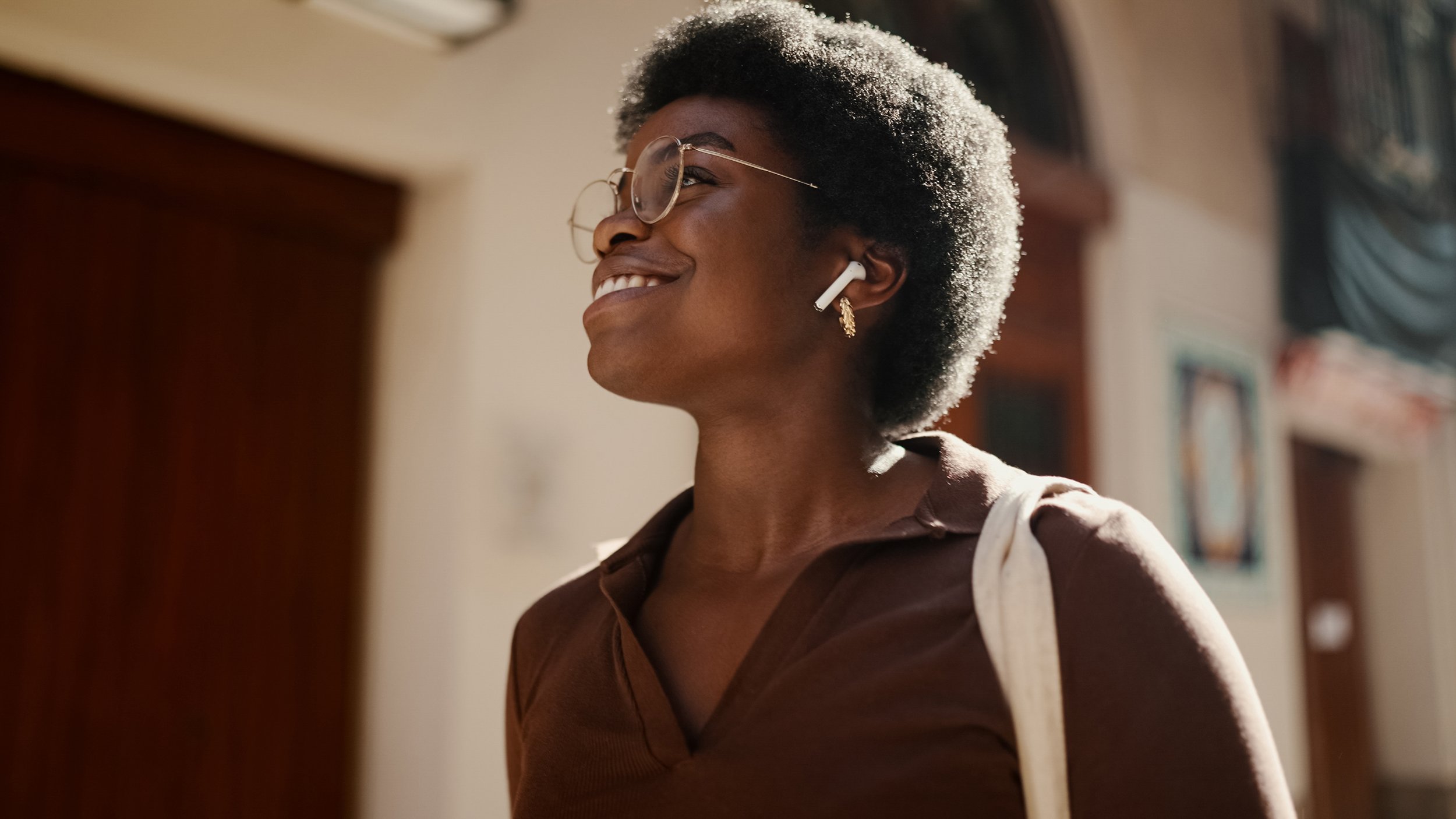 Smiling woman with glasses and wireless earbuds, wearing a brown shirt, standing indoors with a background of wooden furniture and framed pictures.