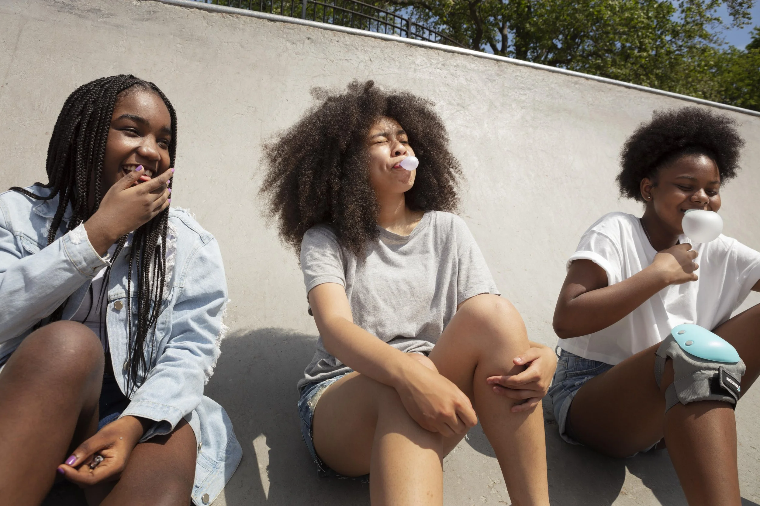 Three young women sitting against a concrete wall outdoors, each blowing bubbles from bubble gum.