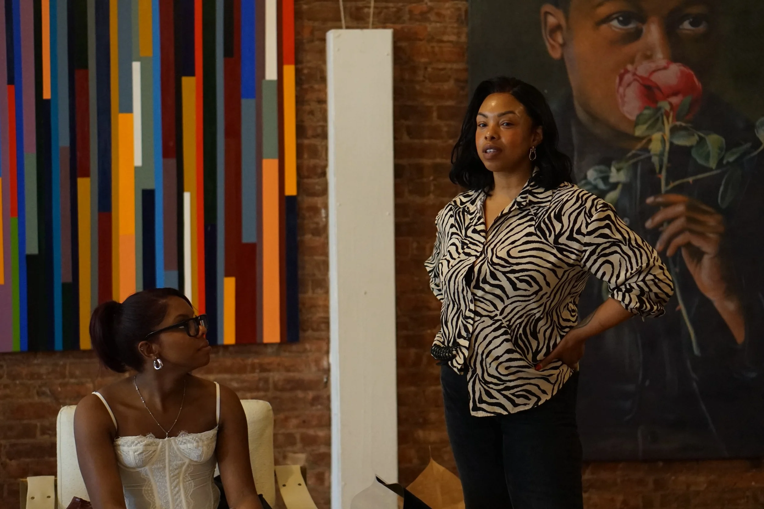 A woman standing and speaking in a room with art on the walls, while another woman seated listens attentively.
