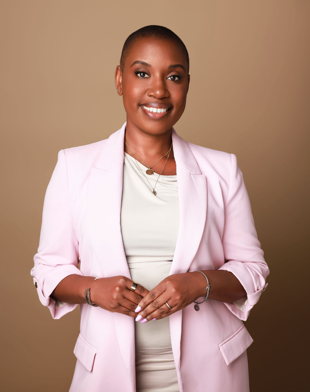 A confident Black woman with a short haircut, wearing a pale pink blazer over a light-colored top, standing against a beige background, smiling with her hands clasped in front of her.
