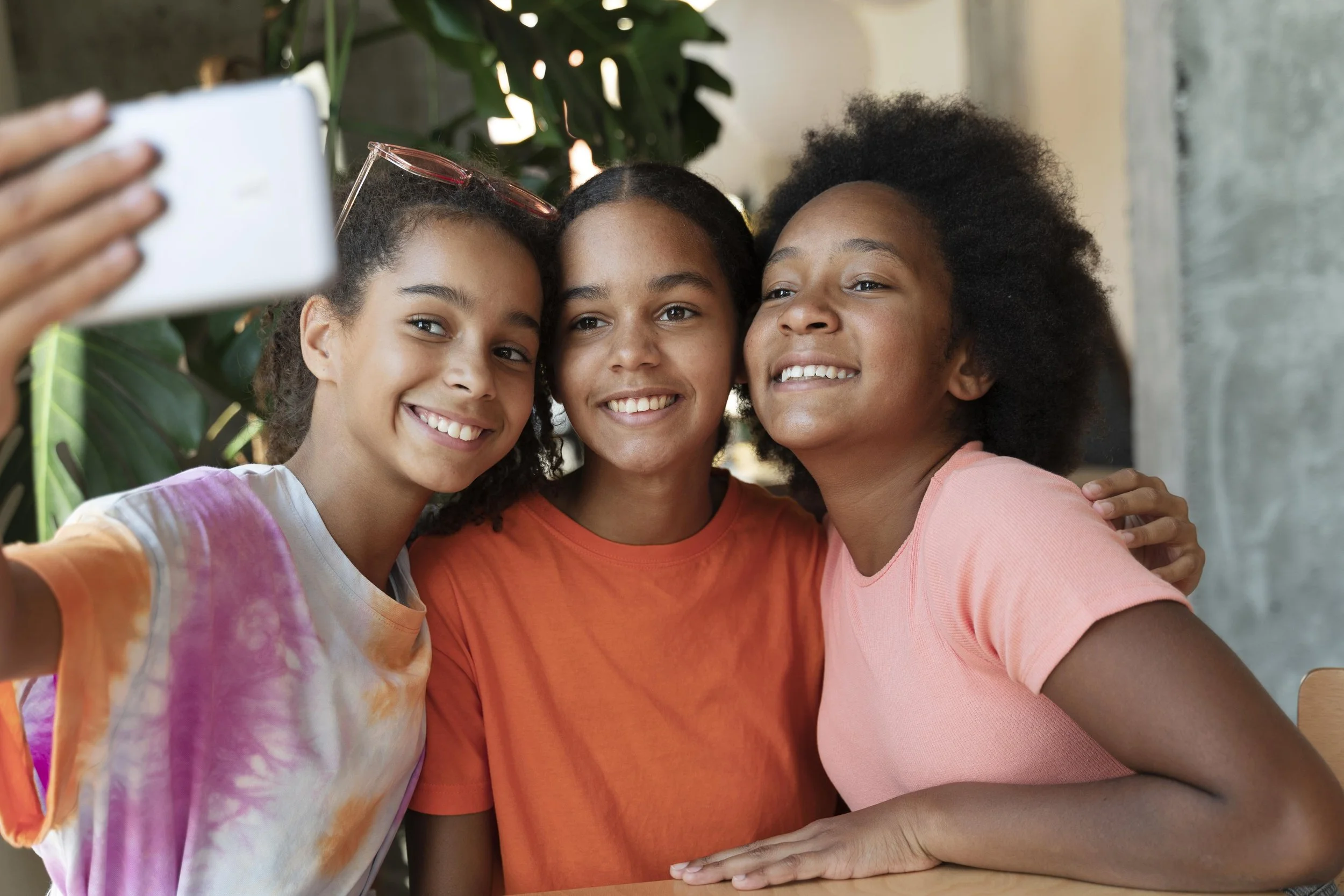 Three smiling young girls taking a selfie together in an indoor setting with greenery in the background.
