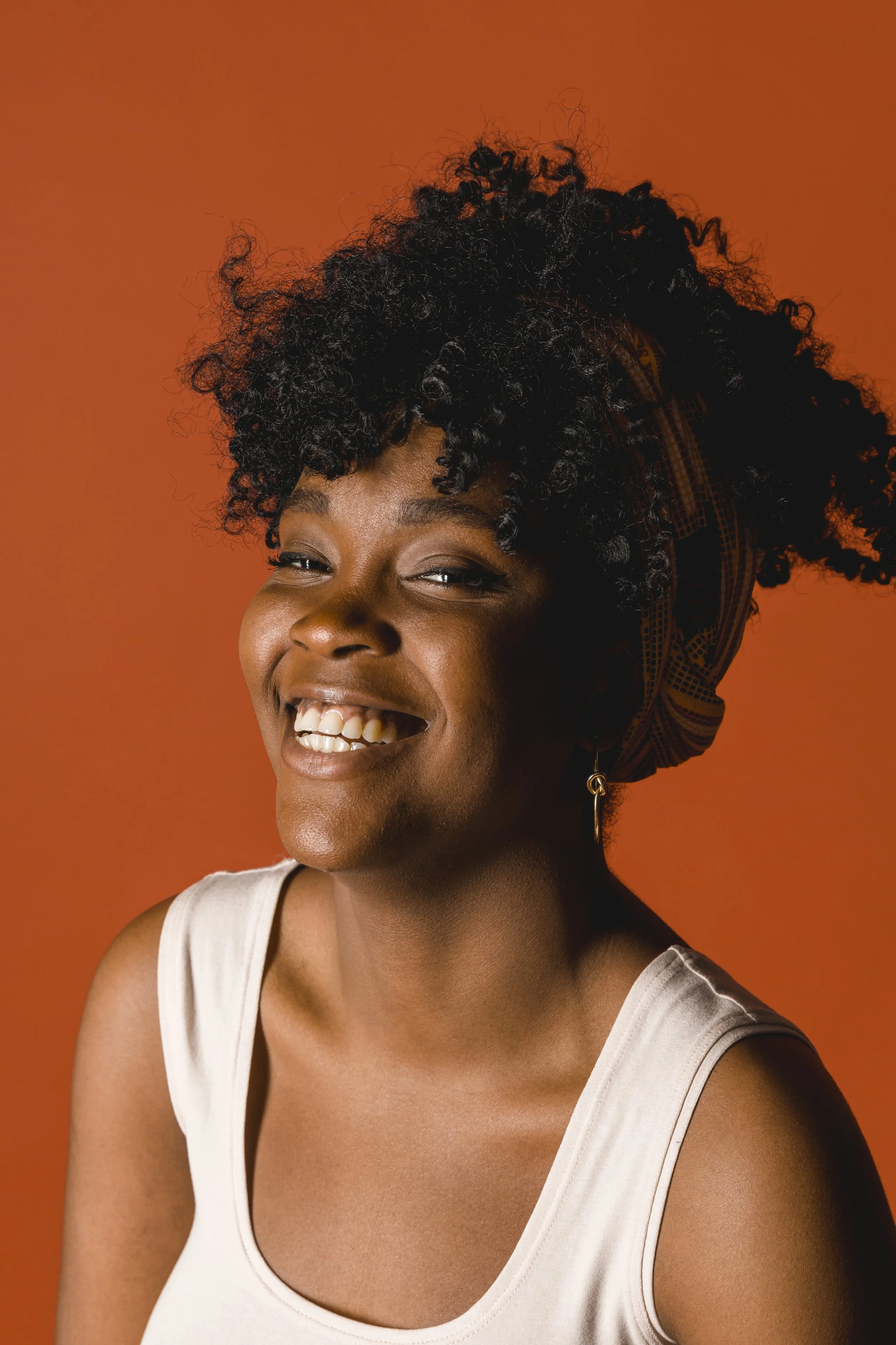 A young woman with dark, curly hair styled with a patterned head scarf, wearing a white sleeveless top, smiling and showing teeth, against an orange background.