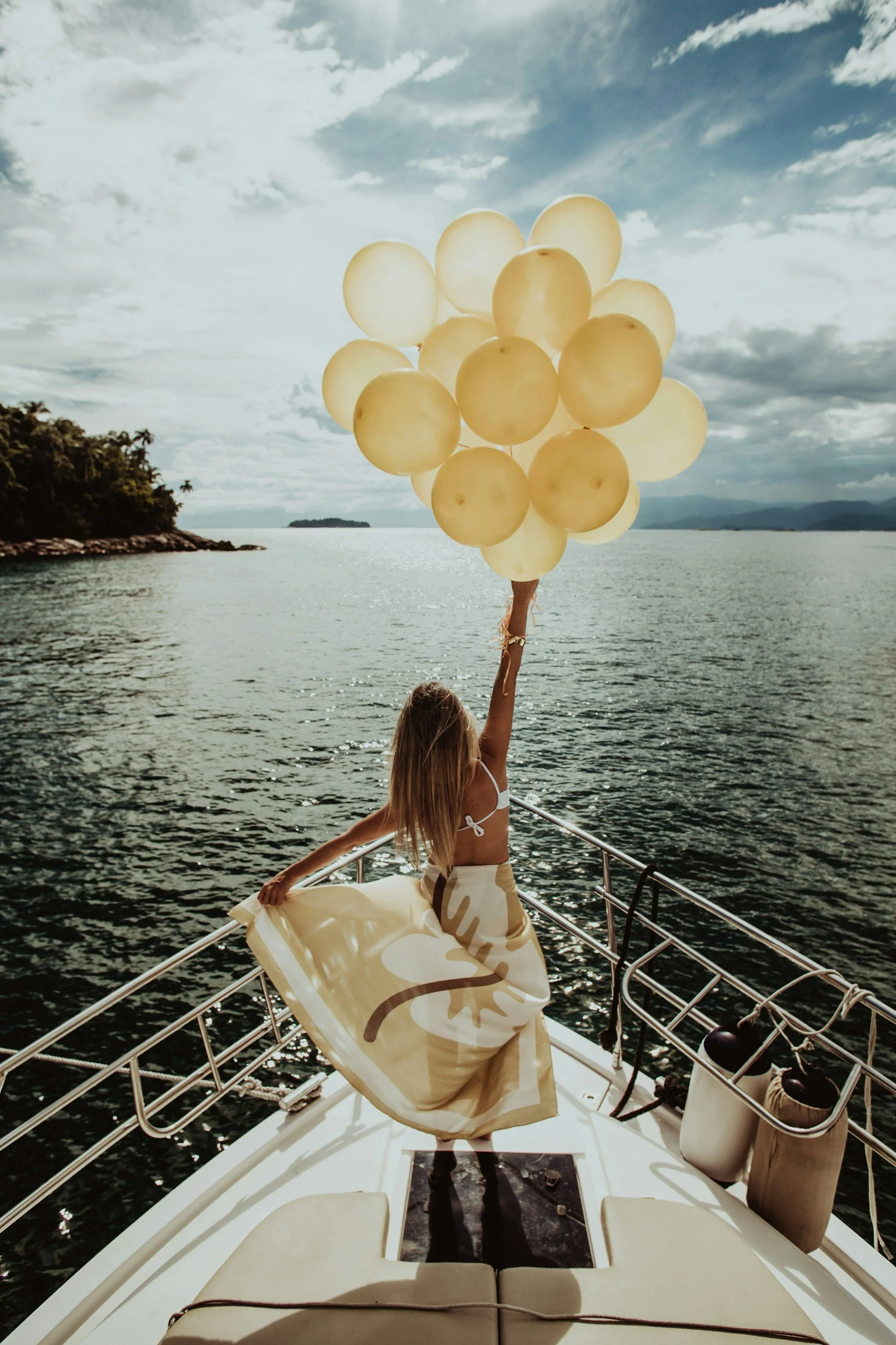 A girl on a boat holding a bunch of light-colored balloons, with an ocean and cloudy sky in the background.