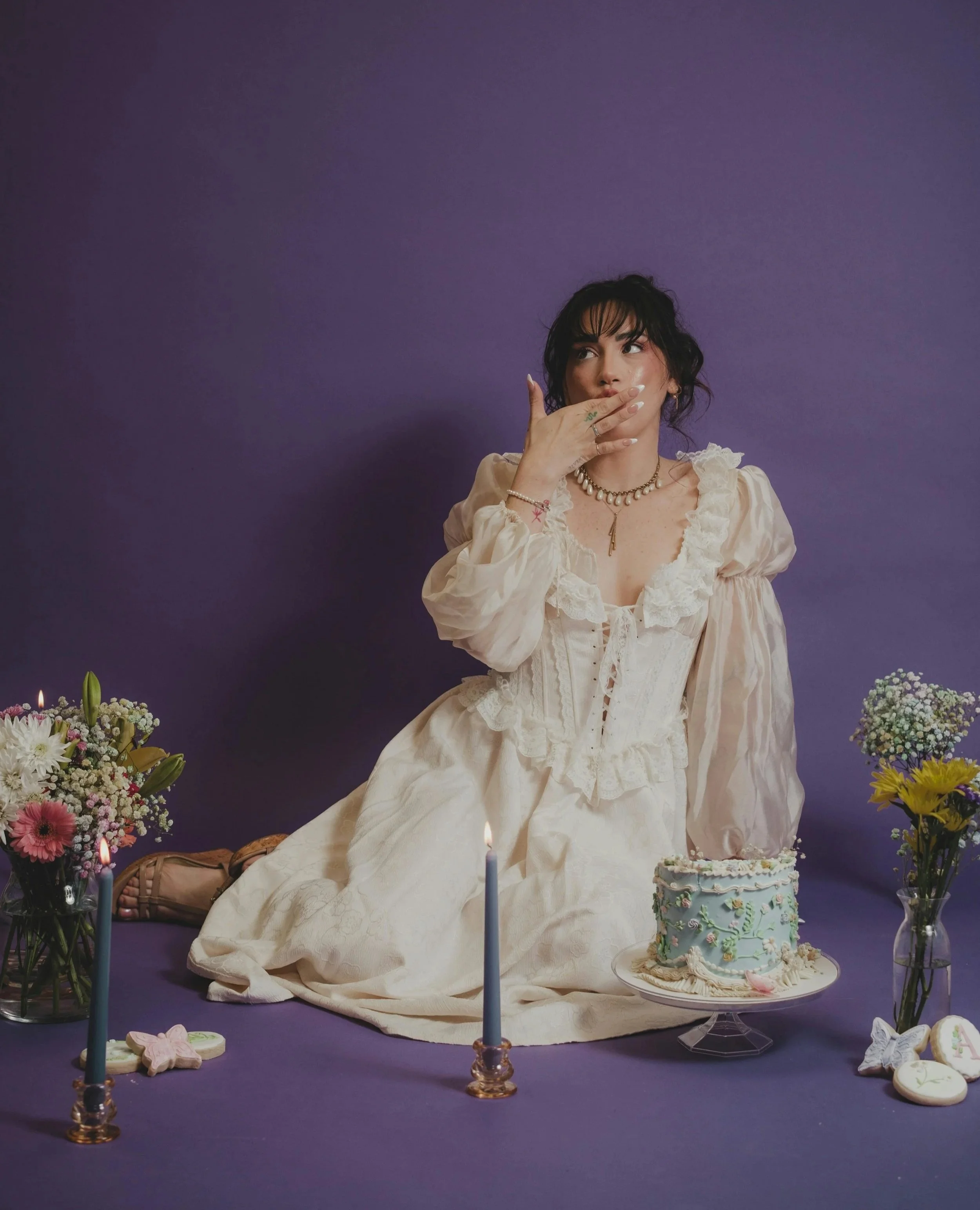 Woman in vintage white dress kneeling on purple background surrounded by flowers, lit candles, and a blue cake with green and pink decorations, holding her hand to her face in surprise.