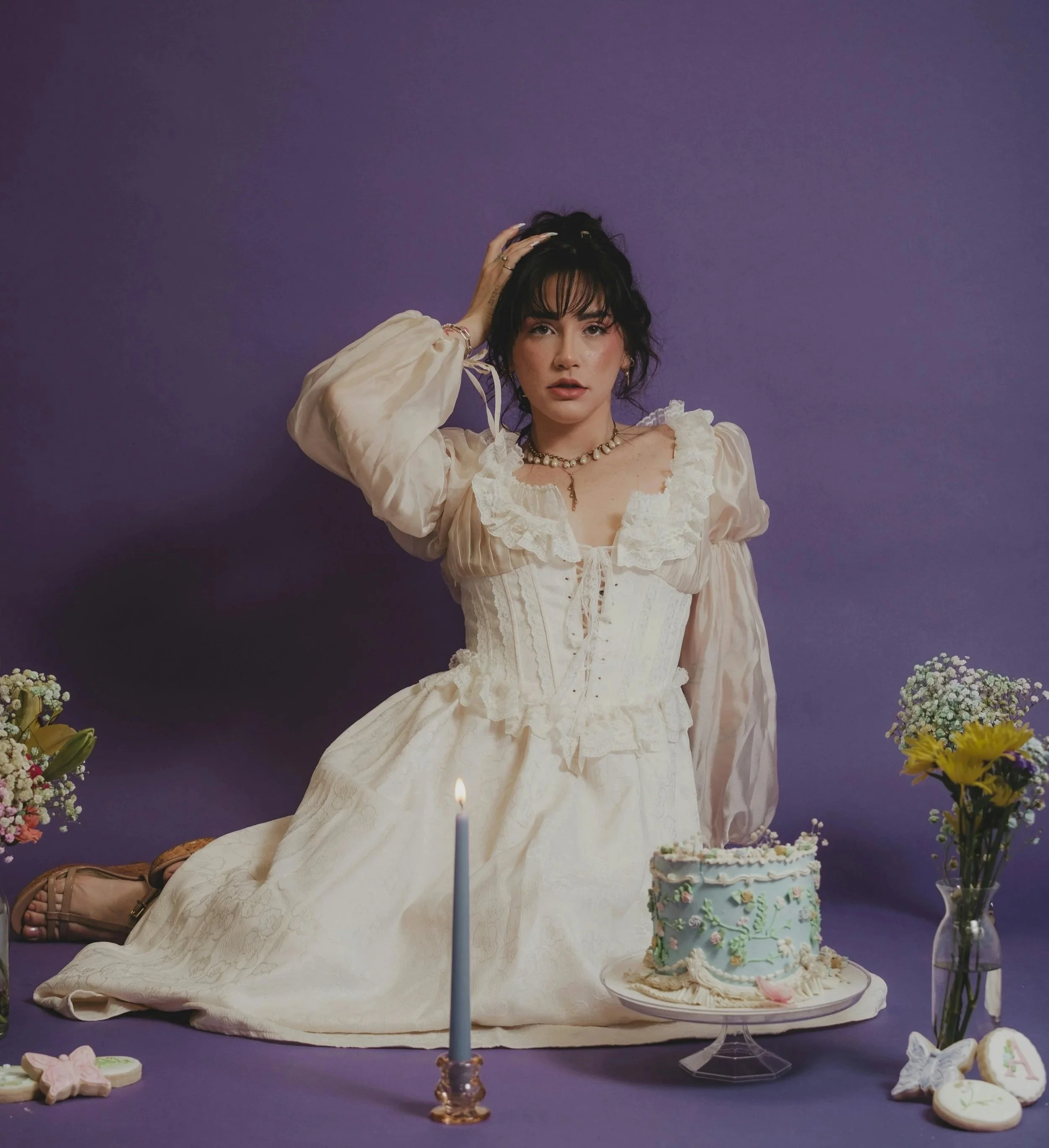 A woman dressed in vintage style white dress with lace details, sitting on the floor next to a decorated cake, surrounded by flowers, with a purple background and a lit candle in front of her.