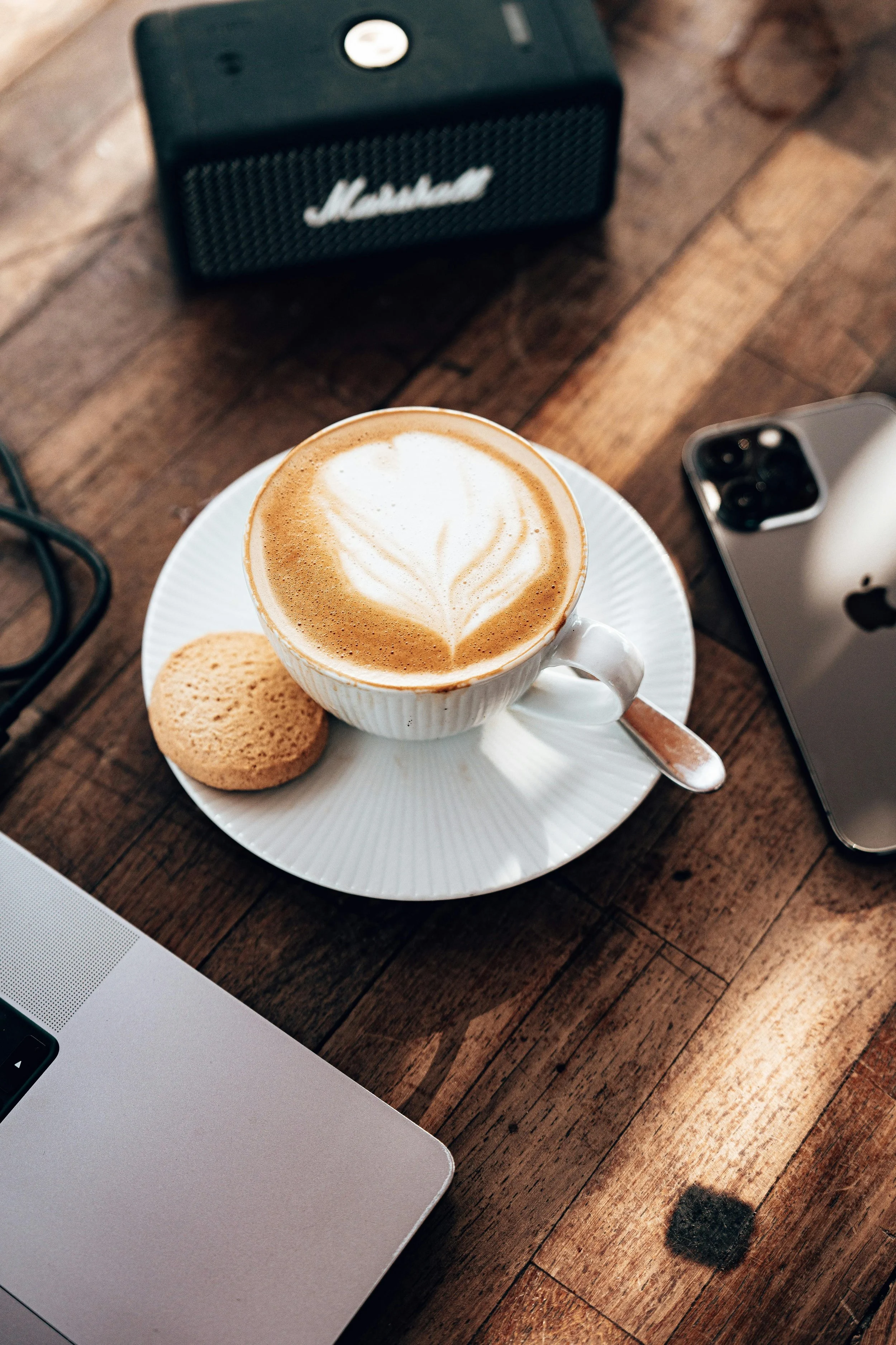 A latte with latte art in a white ceramic cup on a saucer, with a cookie on the side, placed on a wooden table. Also on the table are a smartphone, a laptop, and a small Marshall speaker.