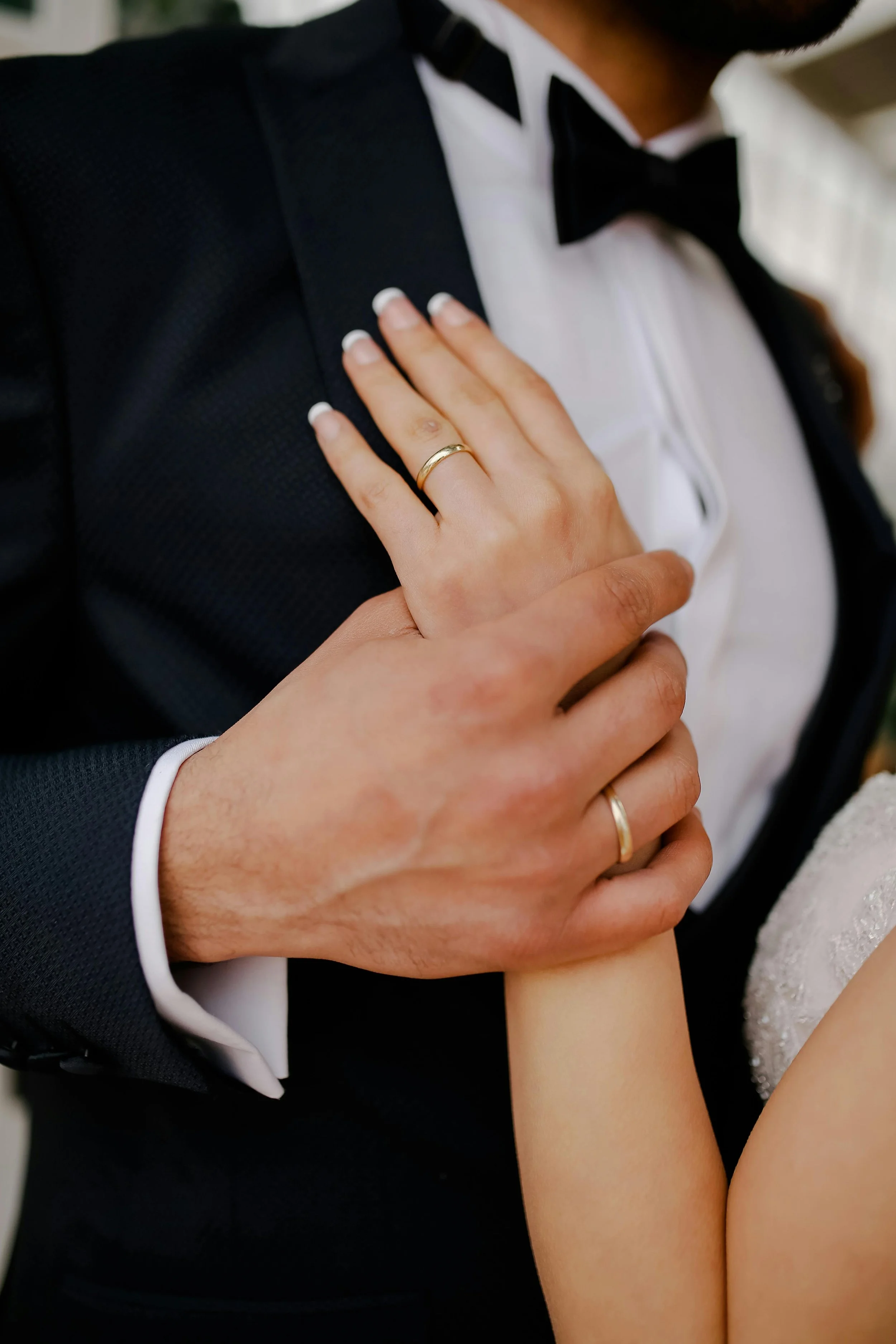 Close-up of a man in a tuxedo with a black bow tie, and a woman with a wedding ring on her left hand resting on his chest.