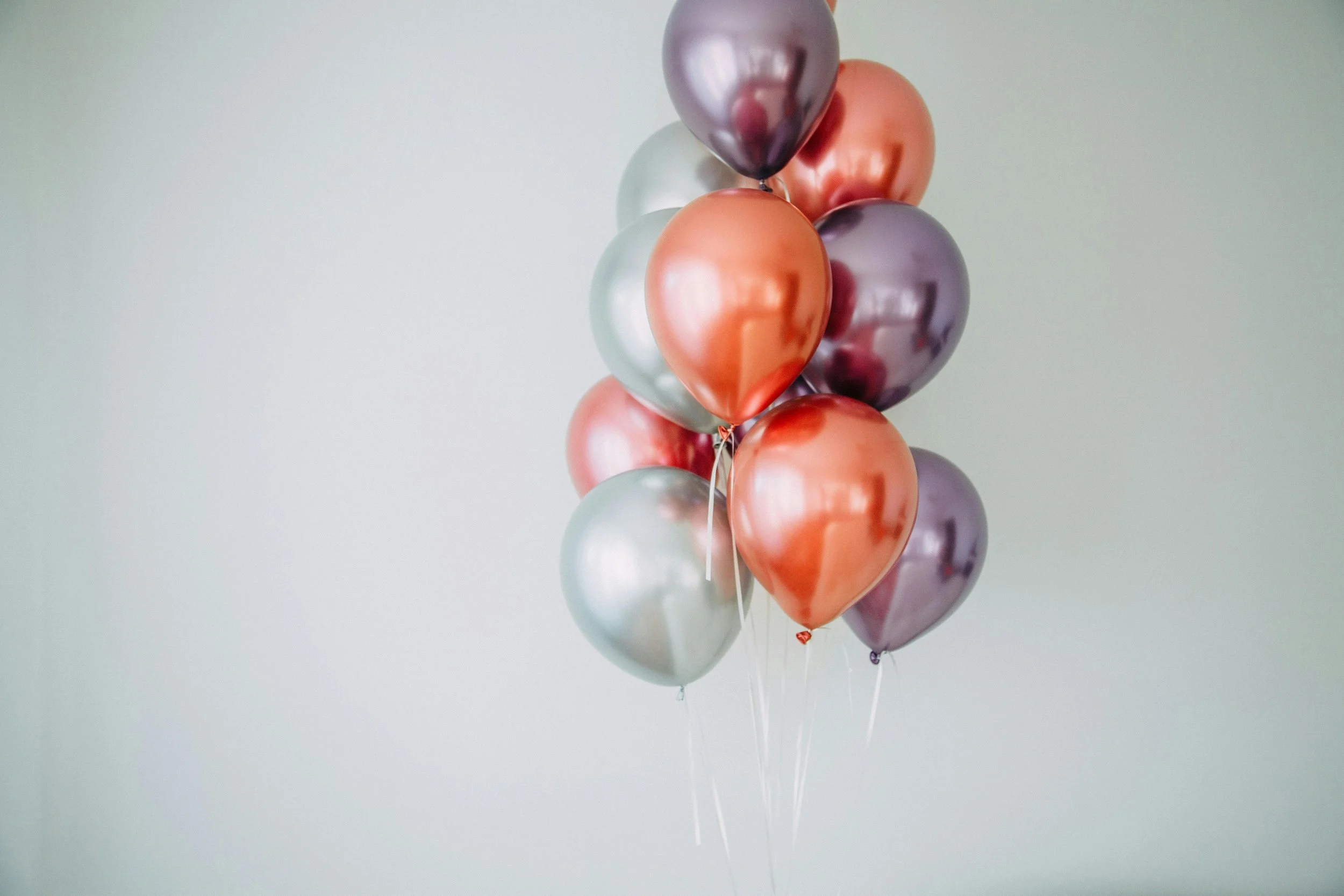 A bunch of shiny metallic balloons in shades of pink, purple, and silver floating against a plain light-colored wall.
