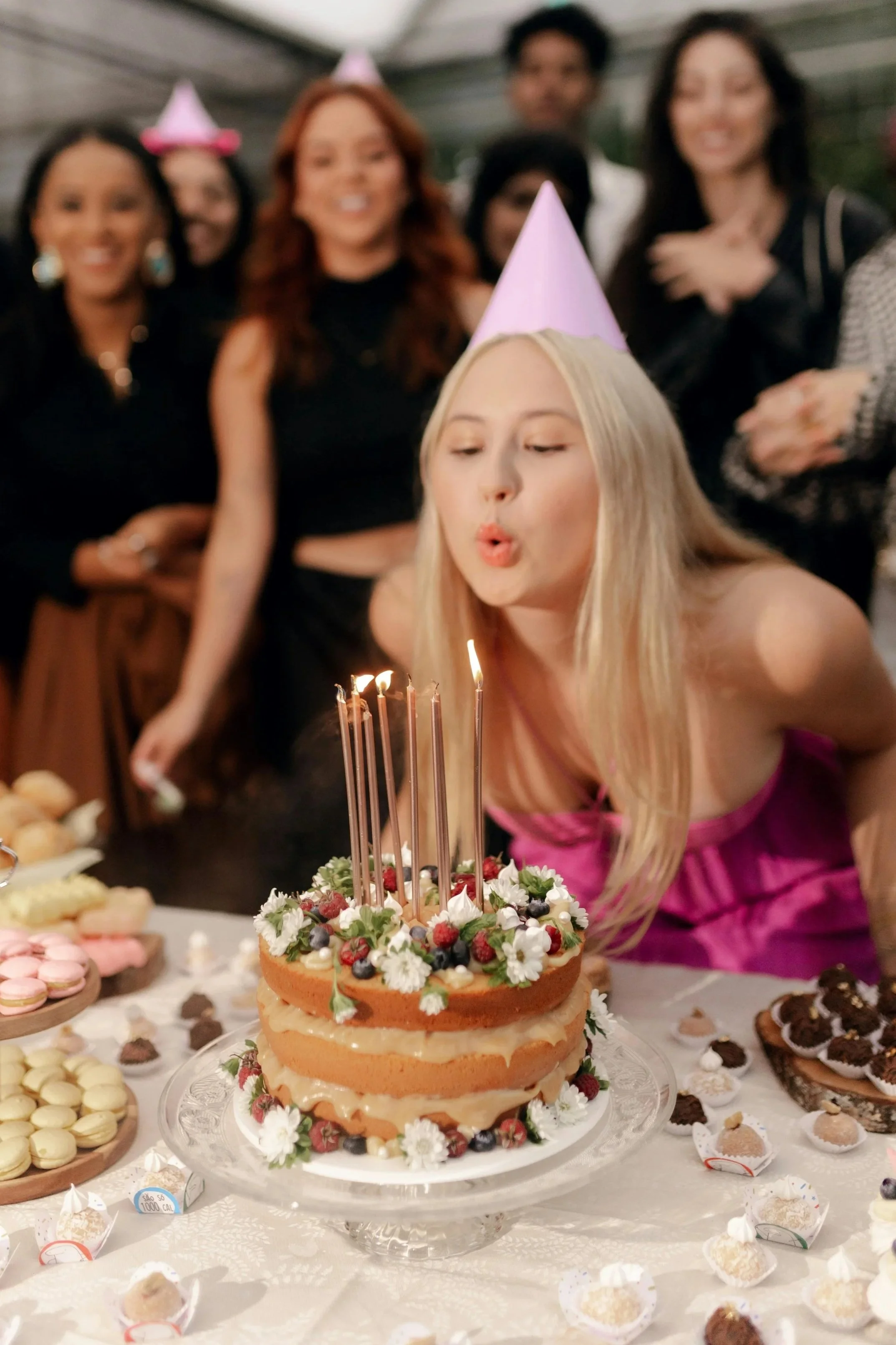 A woman in a pink dress wearing a pink cone birthday hat blows out candles on a layered birthday cake decorated with flowers and berries, with friends in the background wearing pink party hats.