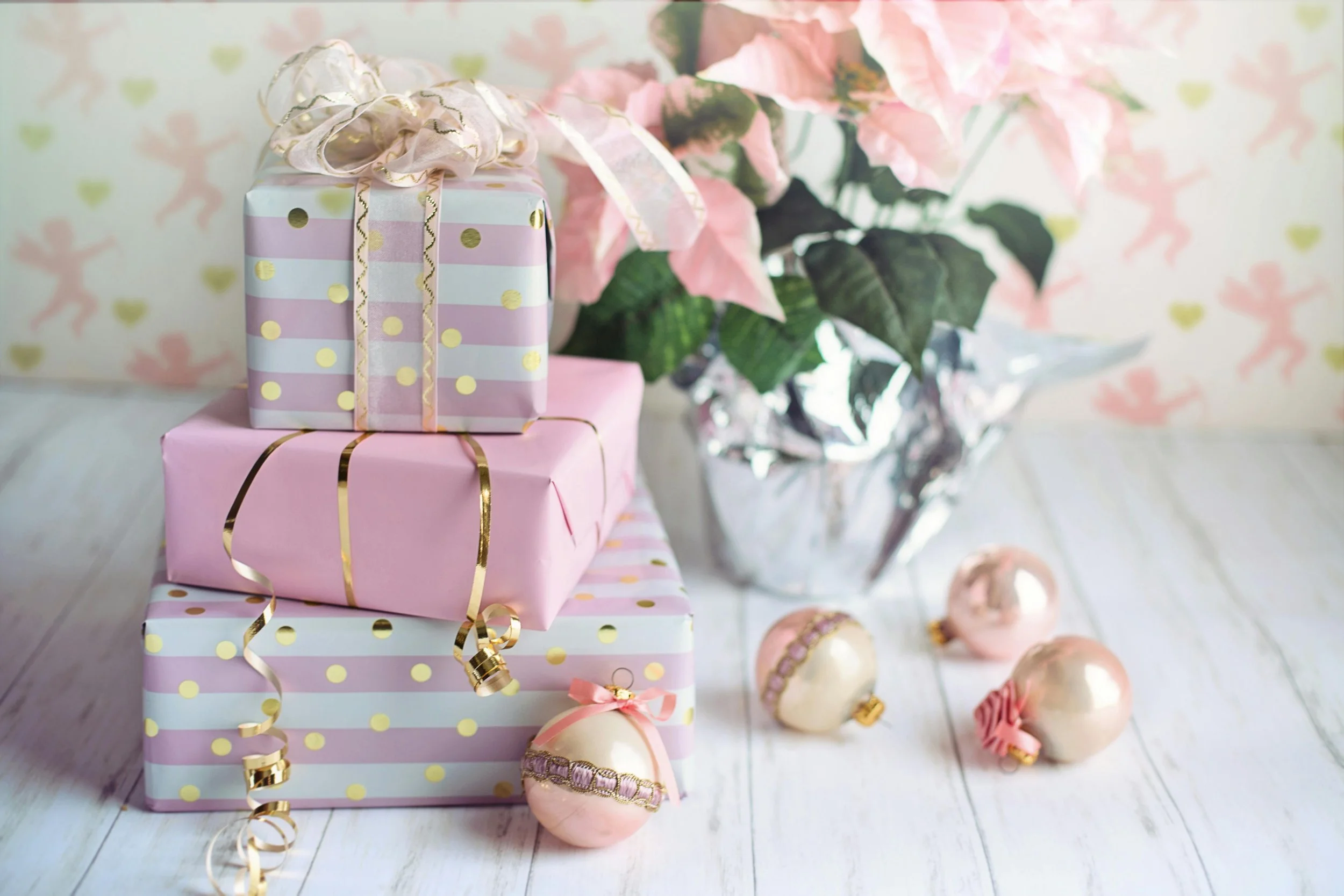 Stacked pastel pink and gift-wrapped boxes with gold accents, decorative ornaments, and poinsettia flowers on a white wooden surface, suggesting a festive holiday setting.