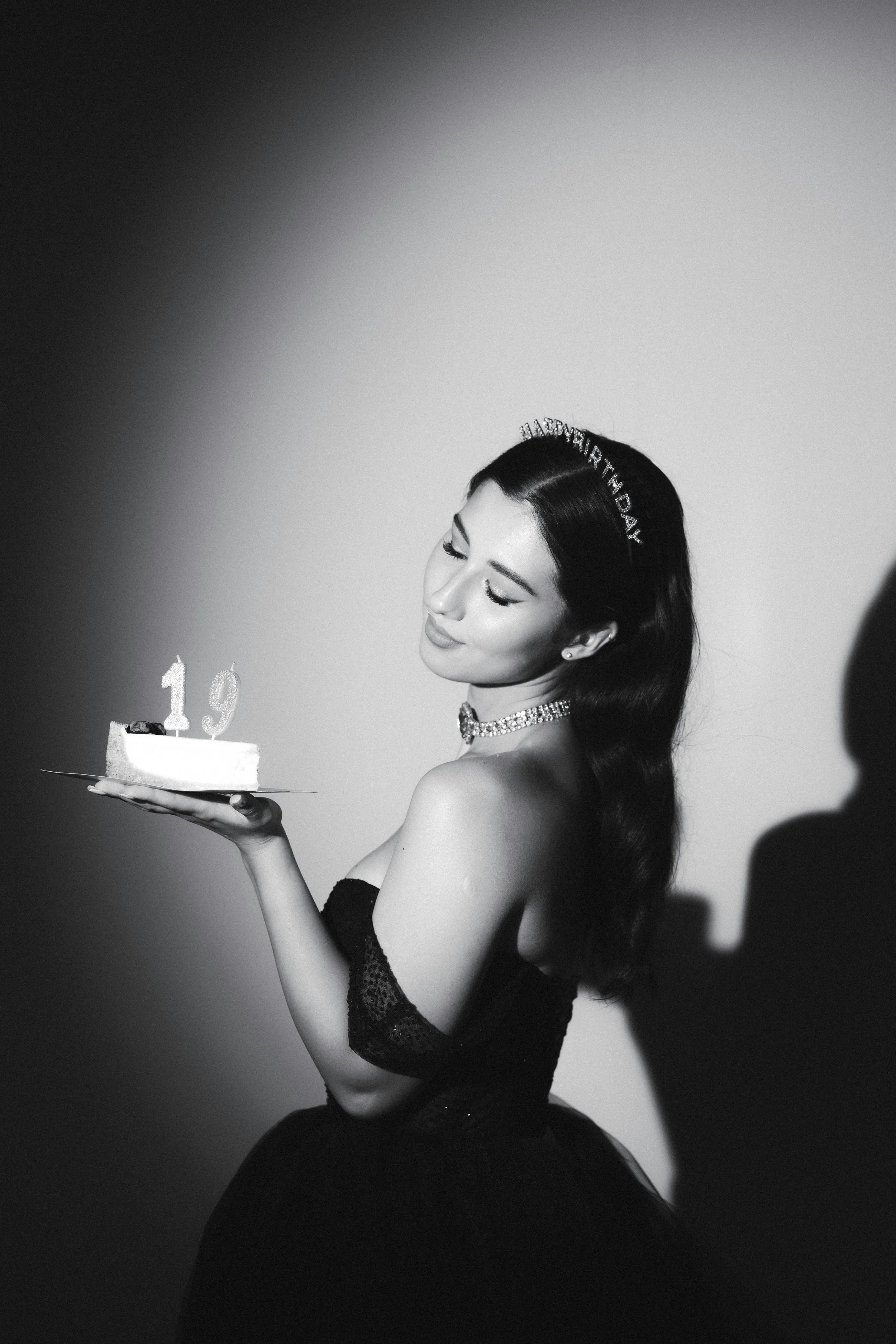 A woman in a strapless dress holding a small birthday cake with candles shaped as the number 19, looking down at the cake, with a shadow cast on the wall behind her.