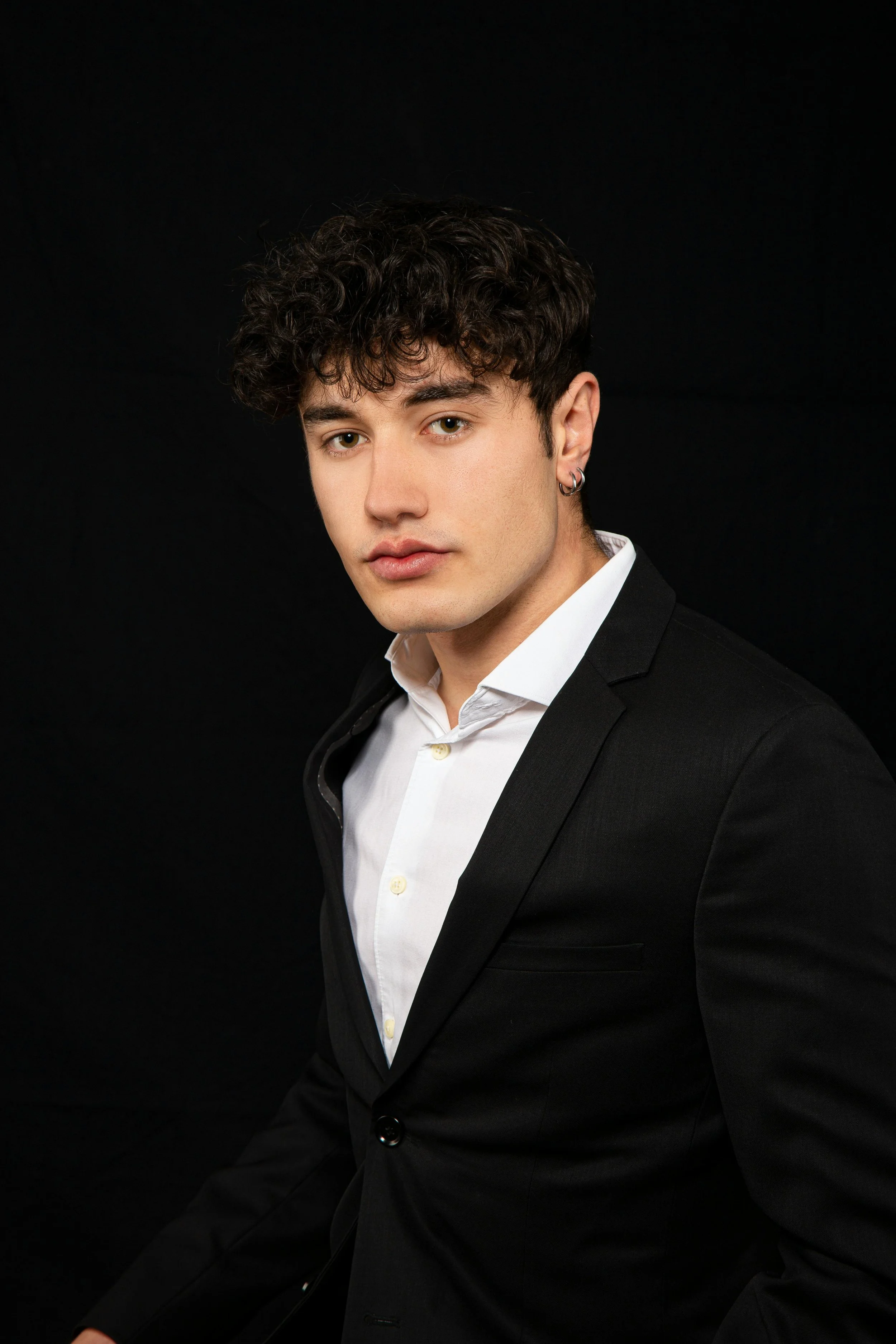 Portrait of a young man with dark curly hair wearing a black suit and white shirt against a black background.