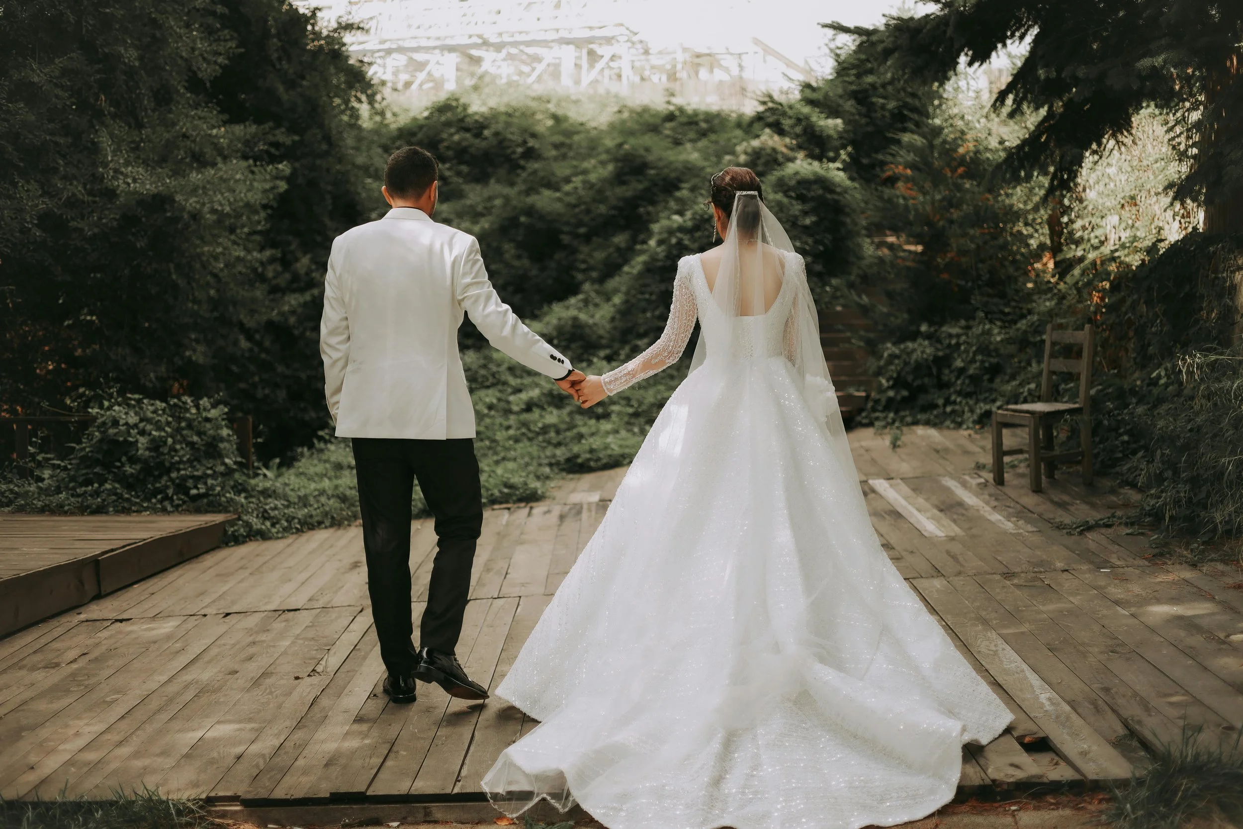A bride and groom holding hands from behind, walking on a wooden platform surrounded by greenery, with an industrial structure in the background.