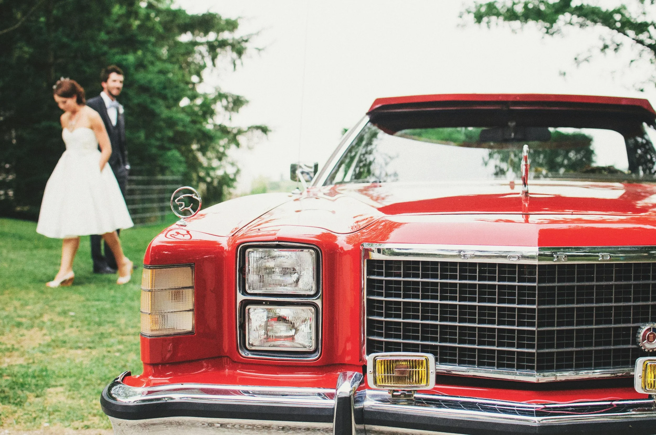 A classic red Ford car outdoors with a couple in wedding attire in the background.