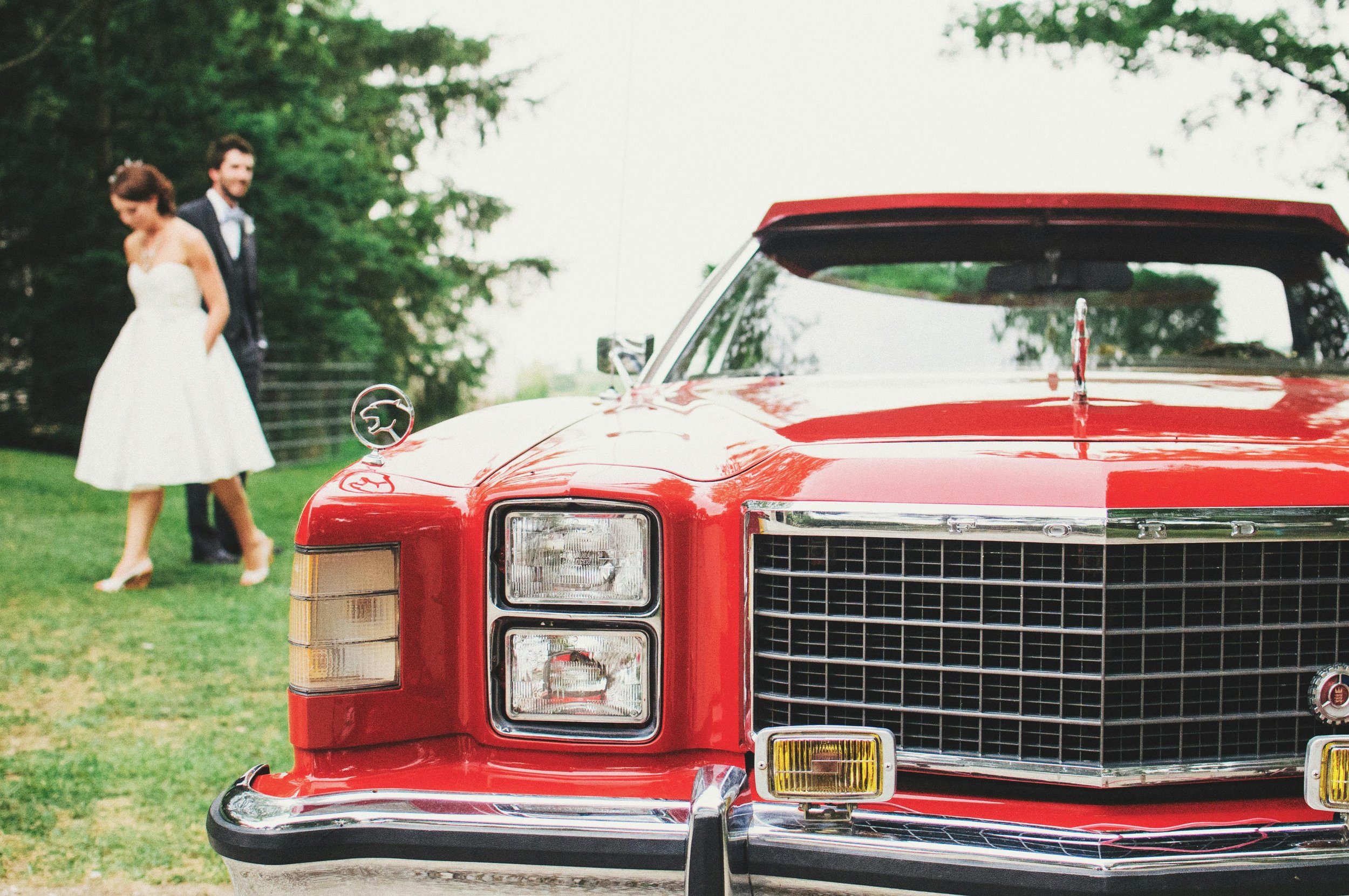 A red vintage Ford car parked on grass with a man and woman in wedding attire standing in the background.